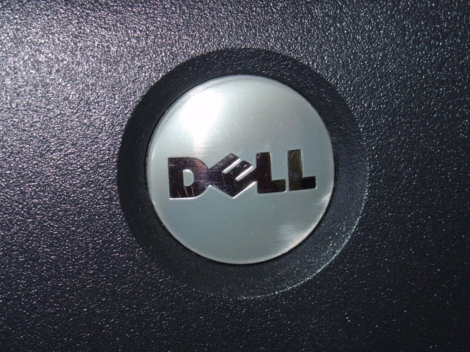 Dell 桌布 壁紙 — 特寫一個銀色 Dell 標誌徽章，設置在帶有反光高光的紋理黑色筆記型電腦蓋上。