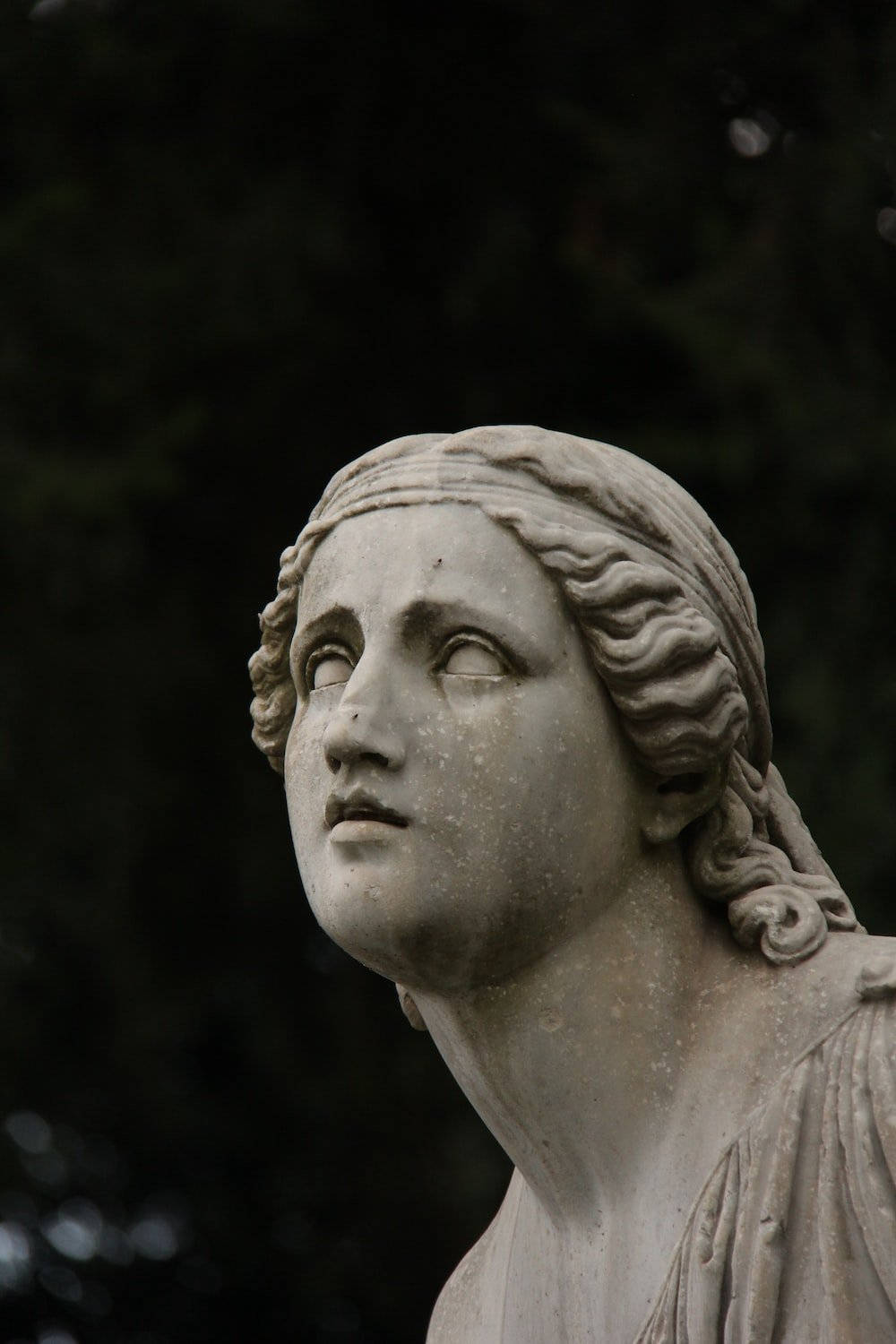 Statue Aesthetic 桌布 壁紙 — 特寫一個風化的古典大理石女性雕像，頭髮飄逸，在黑暗模糊的樹葉背景下向上凝視。