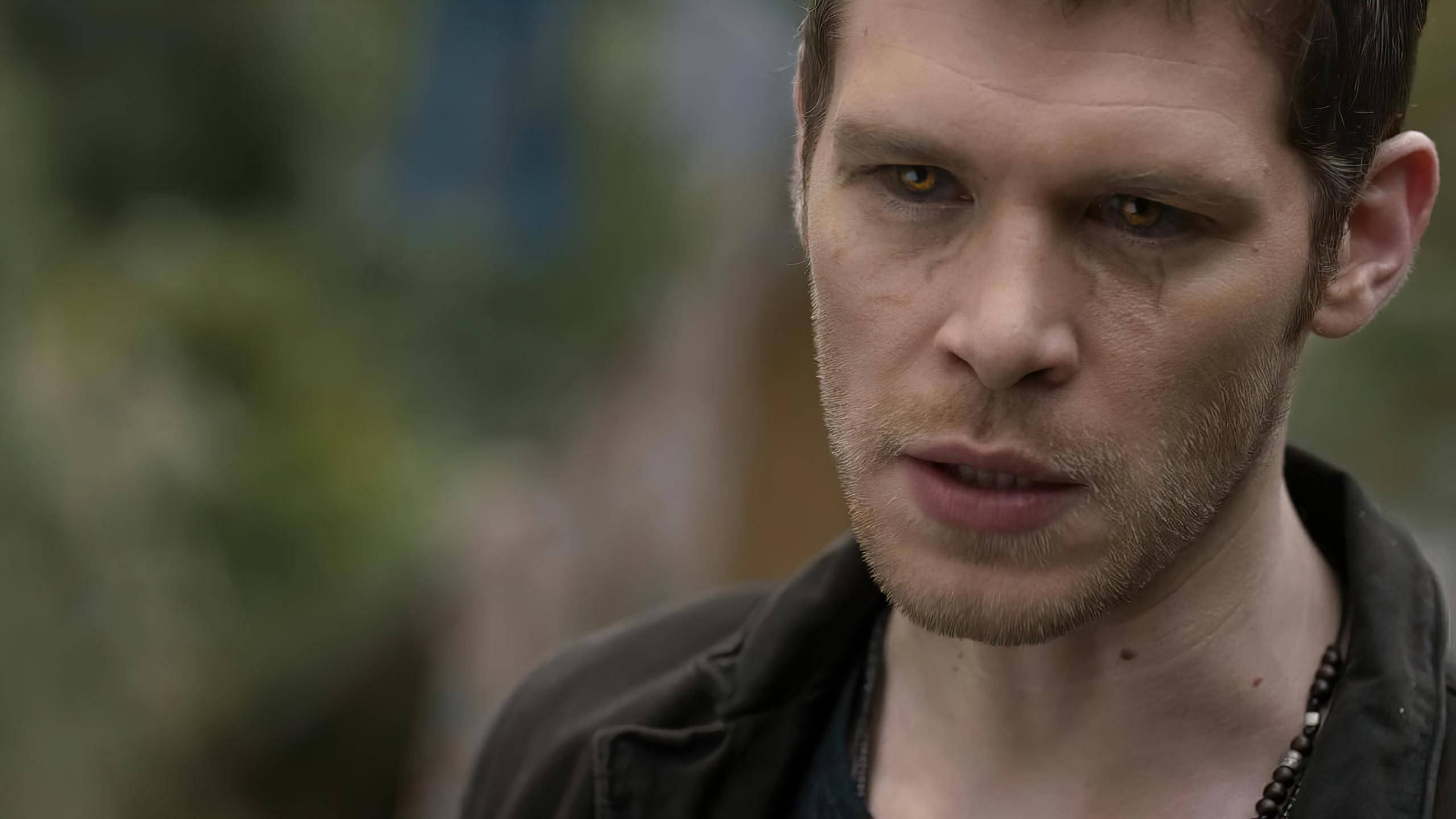 Vampire Diaries 桌布 壁紙 — 特寫一名擁有強烈琥珀色眼睛和鬍渣的男子，穿著深色外套，背景是模糊的自然景色。