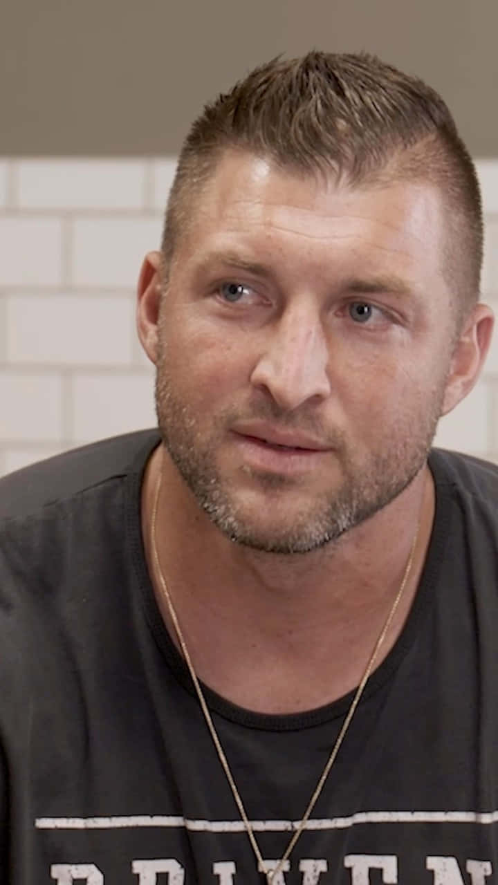 Tim Tebow 桌布 壁紙 — 特寫一名短髮造型、鬍渣明顯的男子，身穿黑色T恤及金鏈，背景為白色磁磚牆。