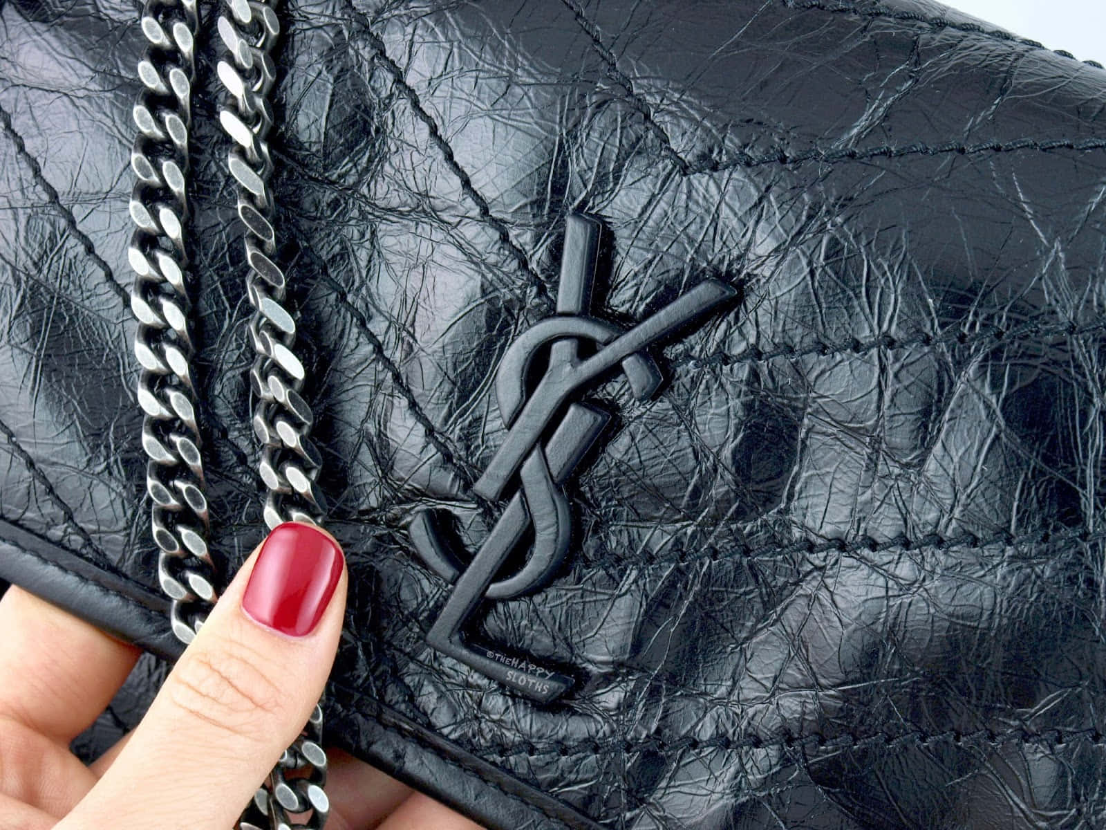 YSL 桌布 壁紙 — 特寫一隻塗有紅色指甲油的手，拿著一個帶有銀色鏈條肩帶的黑色絎縫皮革YSL手袋。