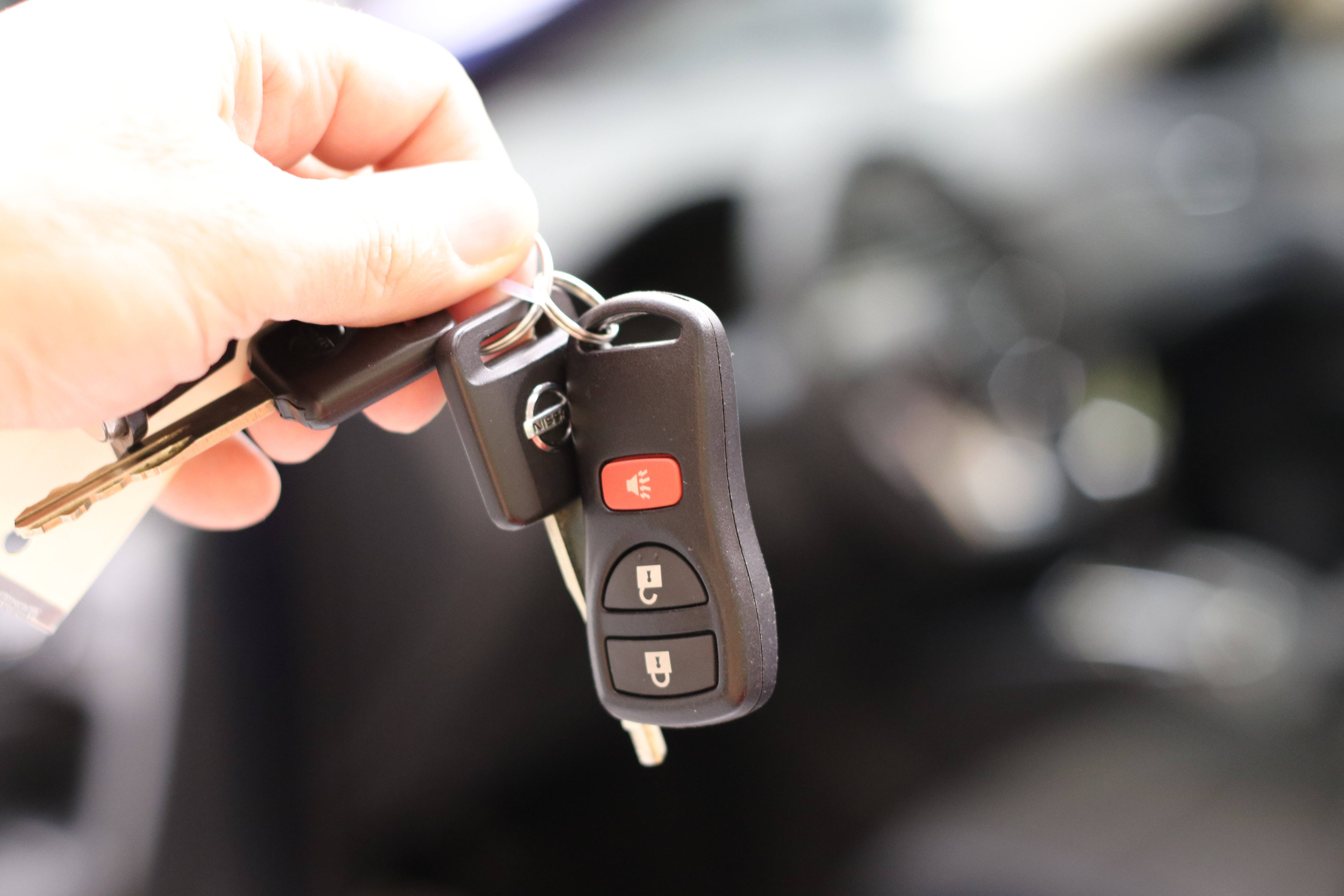 key 桌布 壁紙 — 特寫一隻手拿著一個黑色日產汽車鑰匙扣，帶有紅色緊急按鈕和金屬鑰匙，背景是模糊的車內環境。