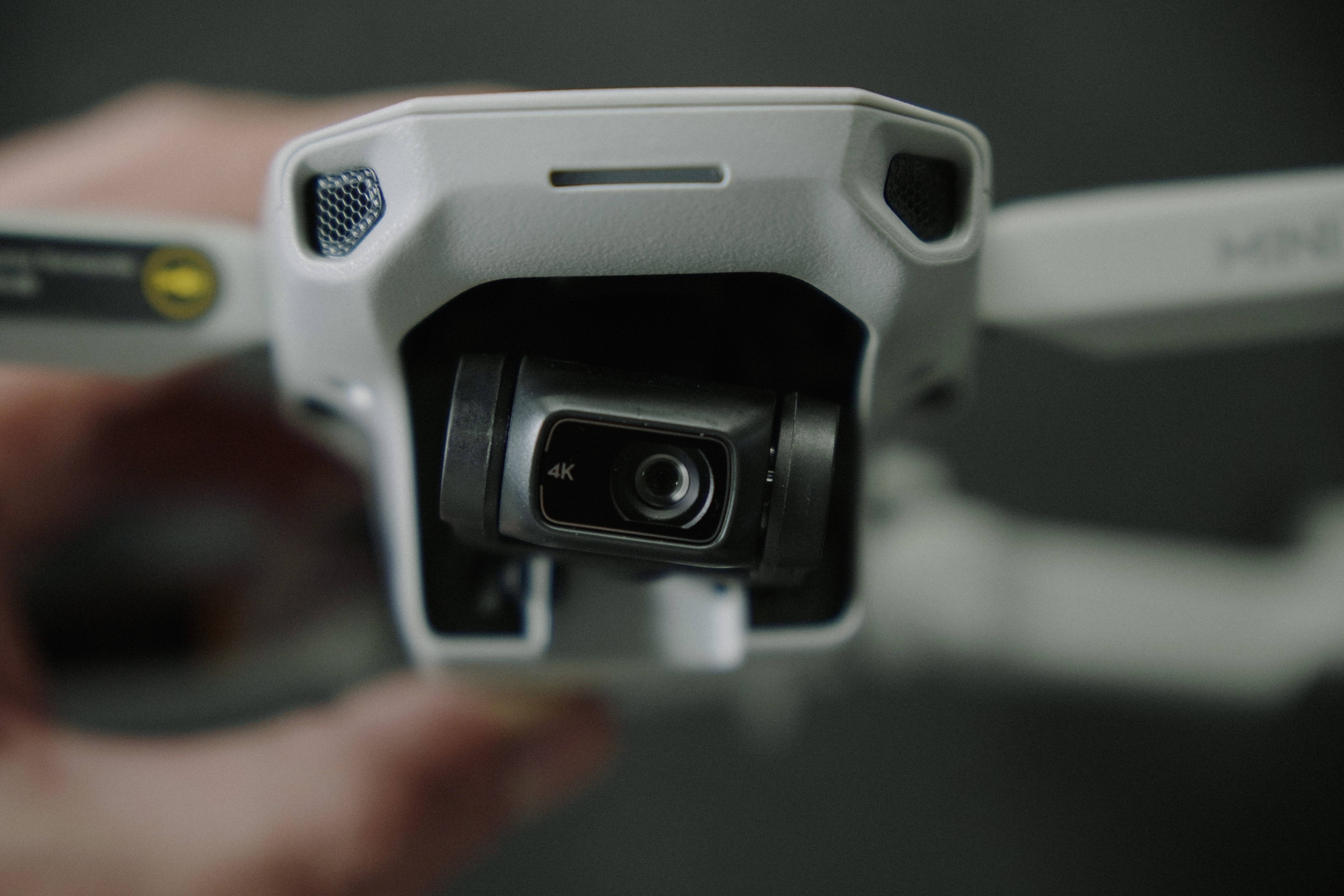 Webcam 桌布 壁紙 — 特寫一隻手拿著一架白色無人機，帶有可見的4K相機鏡頭，背景為深色模糊。