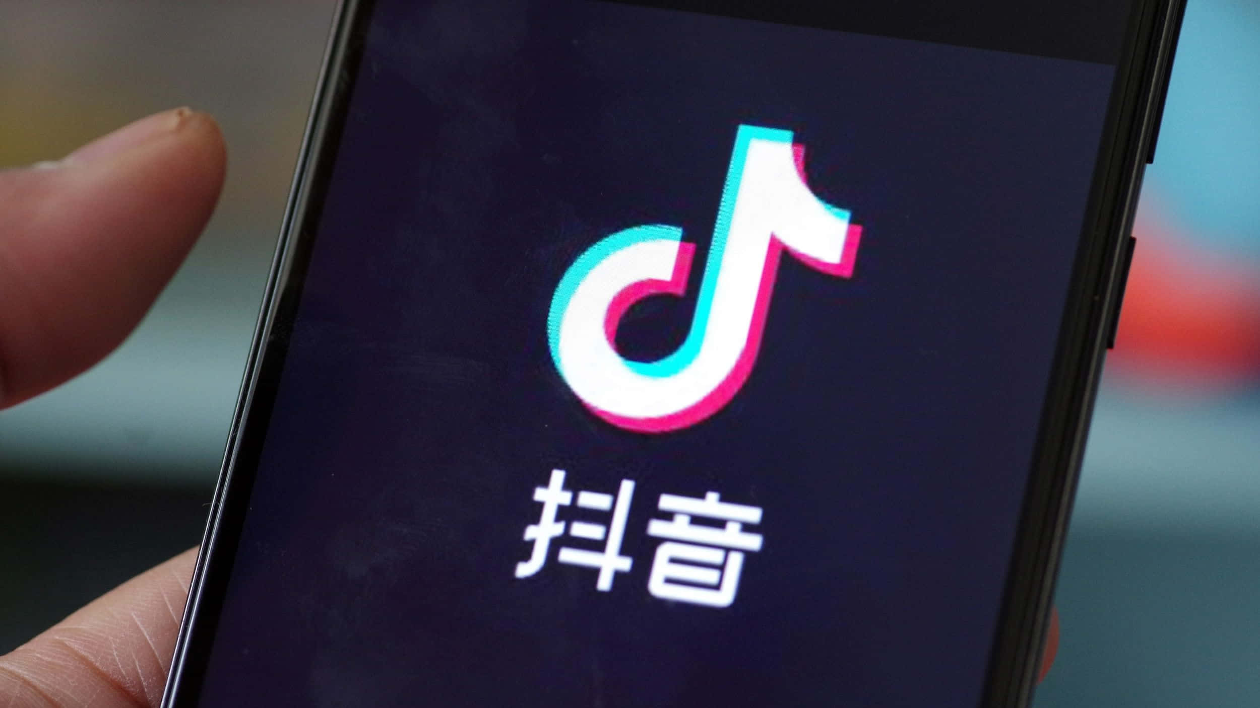 TikTok Logo 桌布 壁紙 — 特寫一隻手拿著智慧型手機，螢幕上顯示色彩繽紛的TikTok標誌，帶有中文字元，背景為深色。