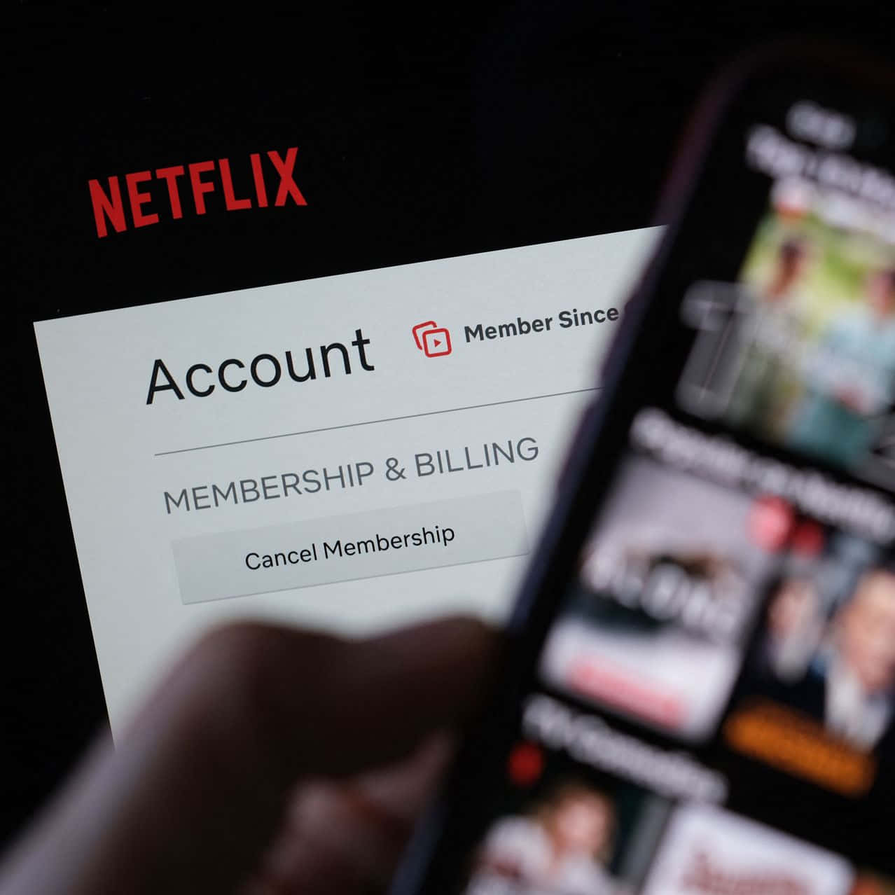 Netflix 桌布 壁紙 — 特寫一隻手拿著智慧型手機顯示Netflix內容，而筆記型電腦螢幕顯示帳戶設定，並有取消會員選項。