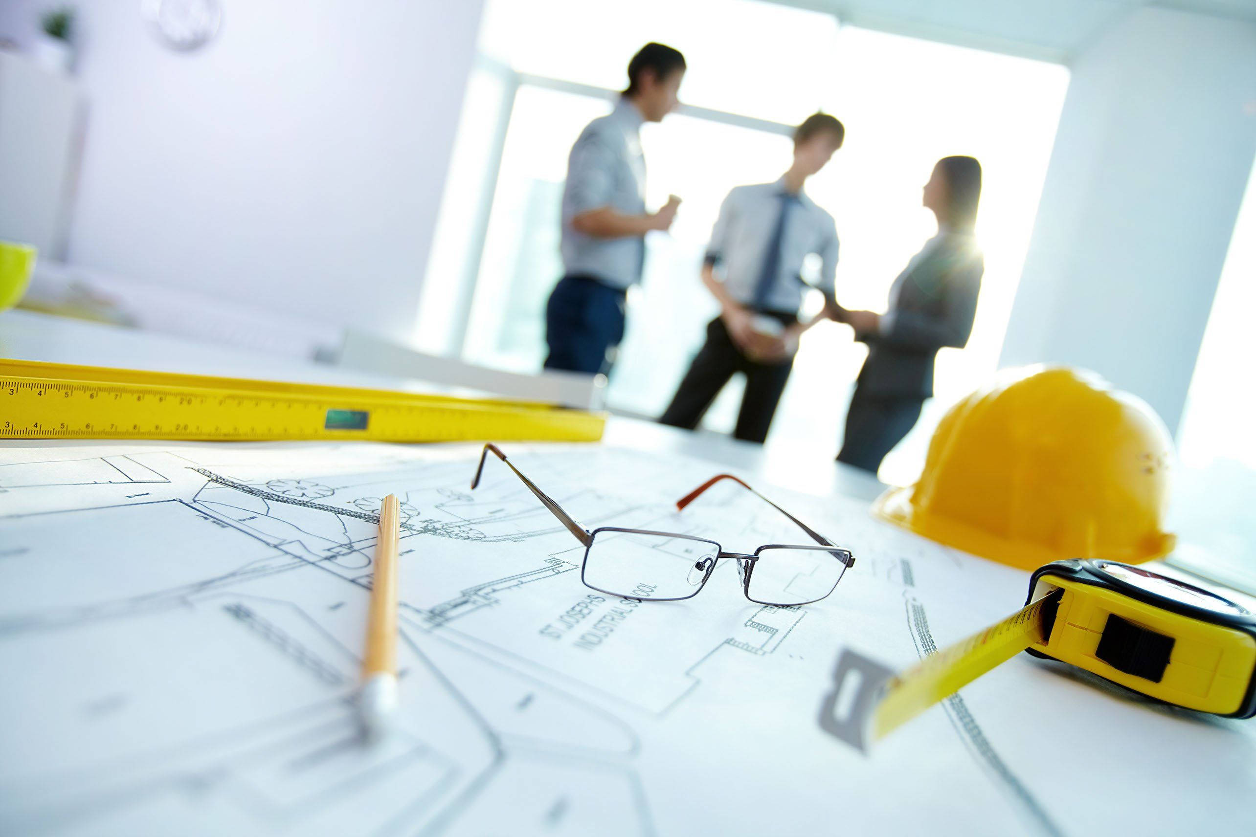 建築 桌布 壁紙 — 特寫建築工具和藍圖在桌上，背景有模糊的專業人士在明亮的辦公室討論。