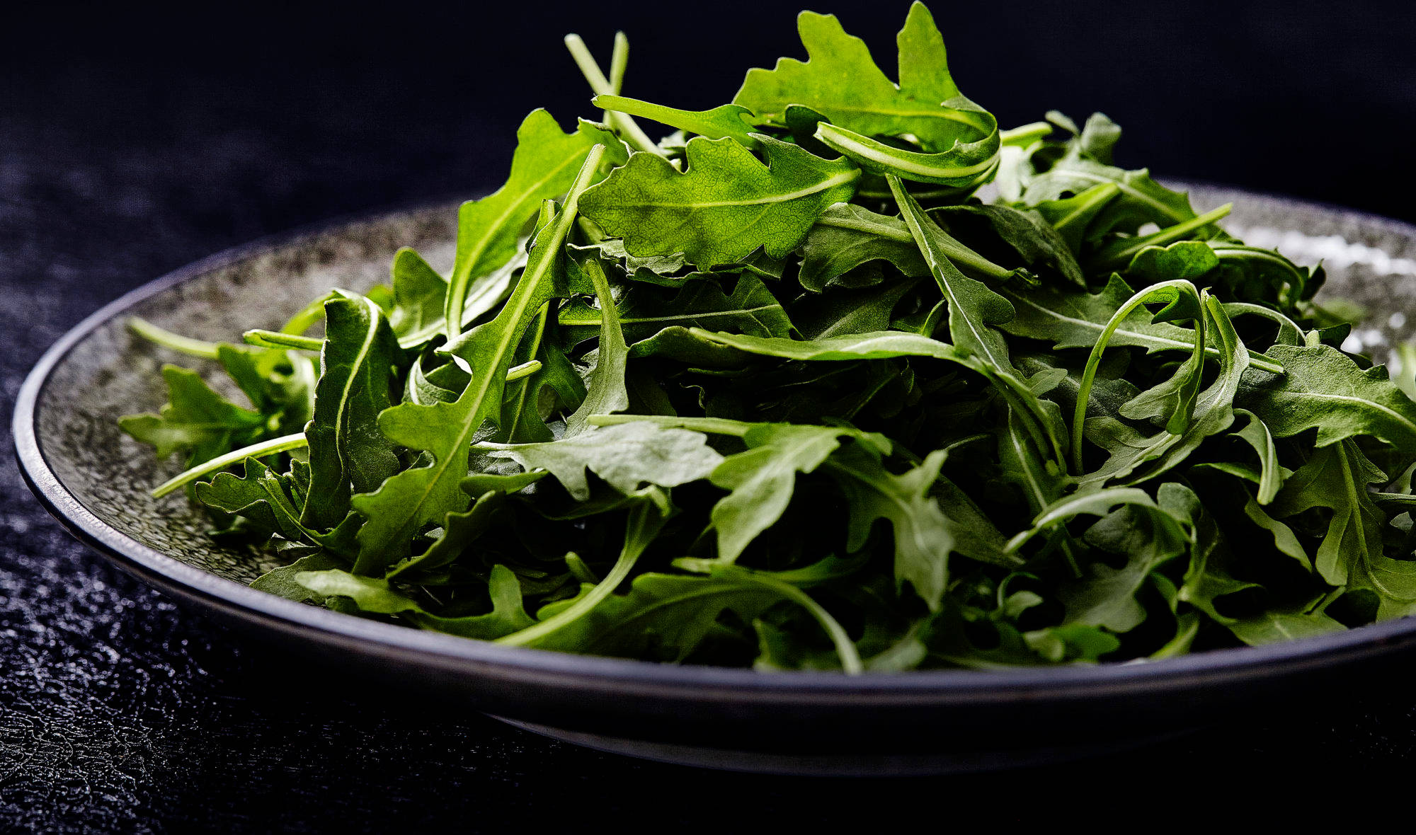Arugula 桌布 壁紙 — 特寫新鮮翠綠的芝麻菜葉，高高堆疊在一個深色紋理陶瓷碗中，背景為黑色。