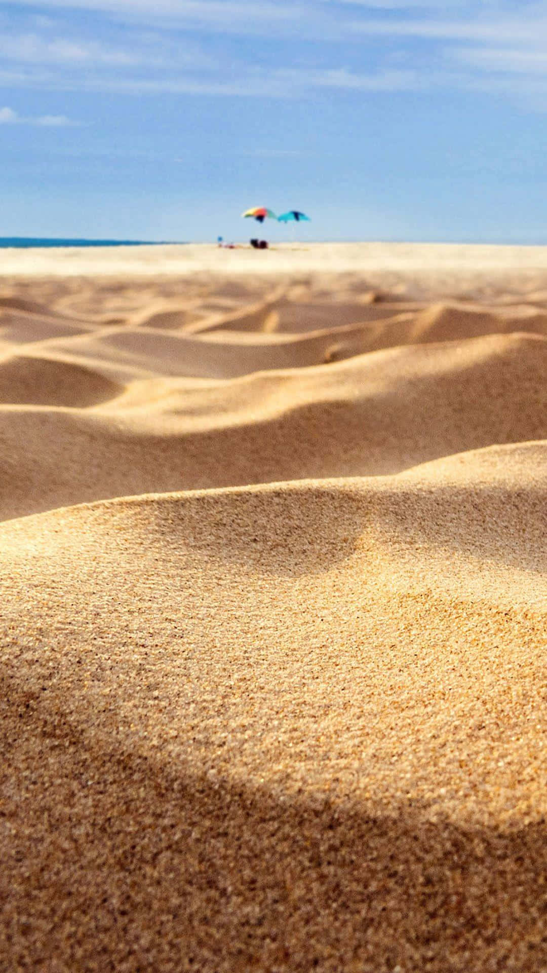 Beach Sand 桌布 壁紙 — 特寫紋理金色沙丘，在明亮的藍天下，遠處有沙灘傘和海洋。