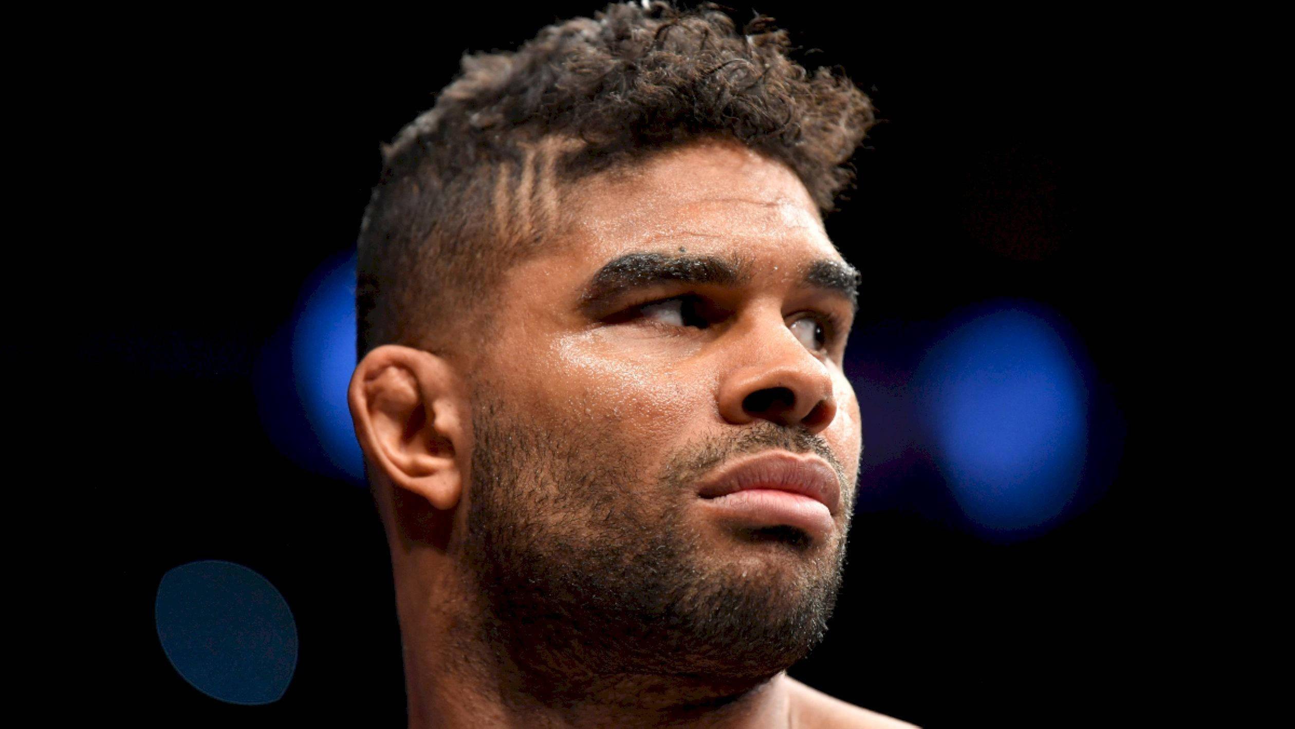 UFC 桌布 壁紙 — 特寫肖像，一位專注的男性戰士，捲髮和鬍鬚，背景為深色帶有藍色散景燈光。