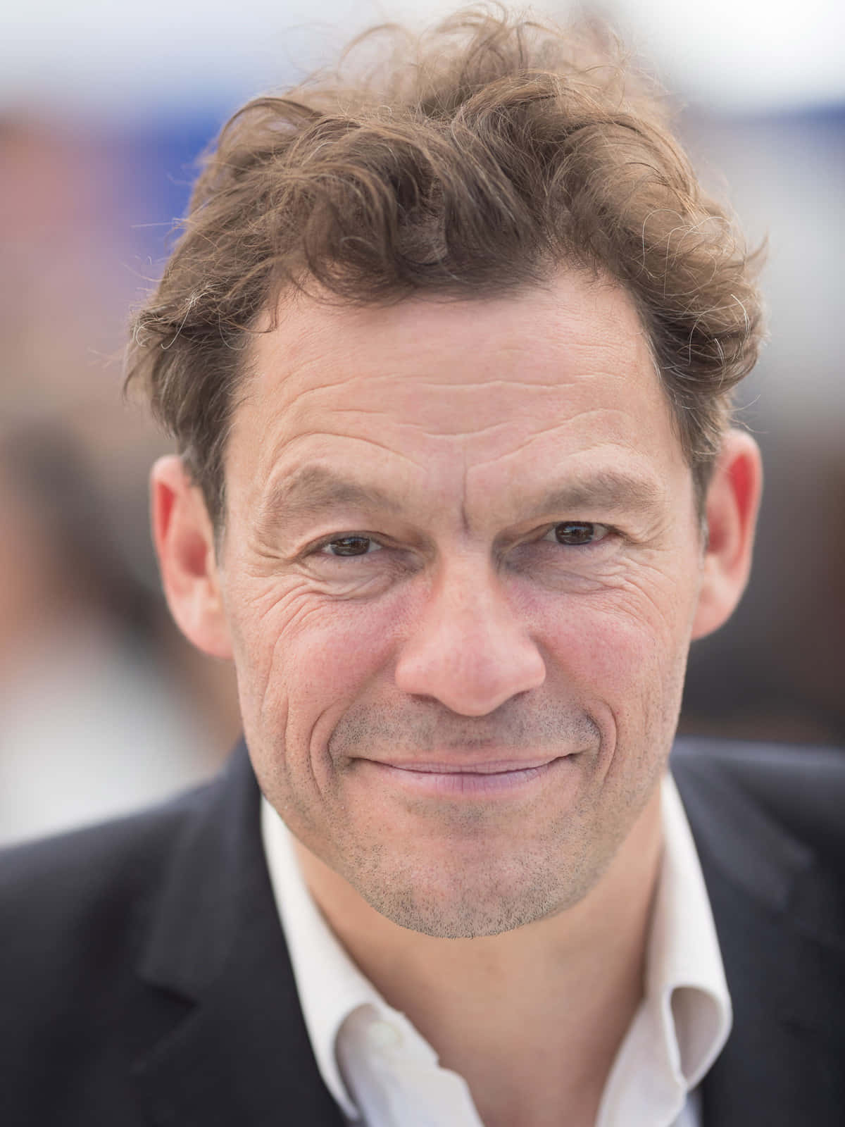 Dominic West 桌布 壁紙 — 特寫肖像，一位微笑的中年男子，一頭凌亂的棕色頭髮，身穿深色西裝，背景模糊。