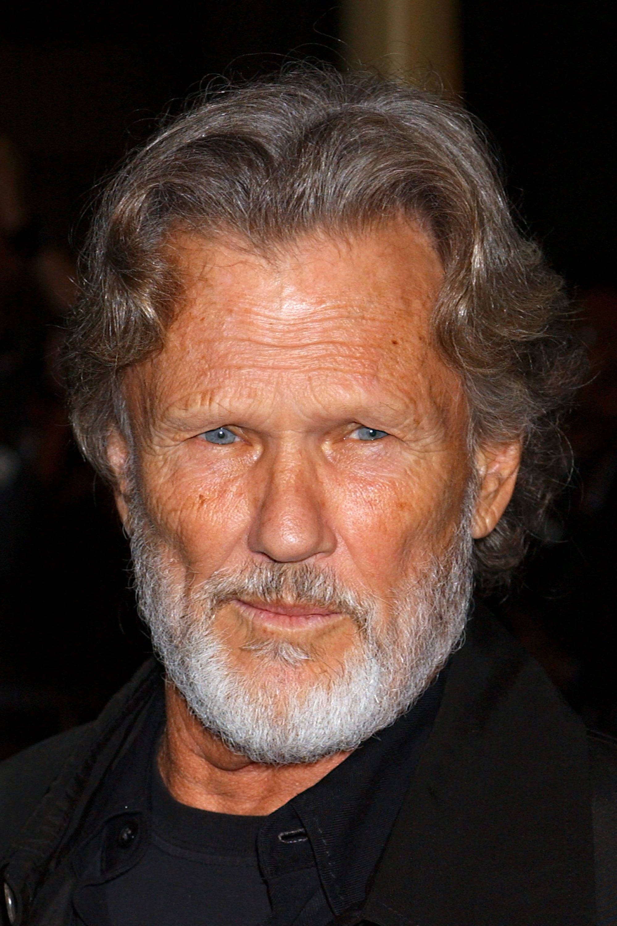 Kris Kristofferson 桌布 壁紙 — 特寫肖像，一位飽經風霜的男子，灰白頭髮和鬍鬚，身穿黑色衣服，背景為深色模糊。