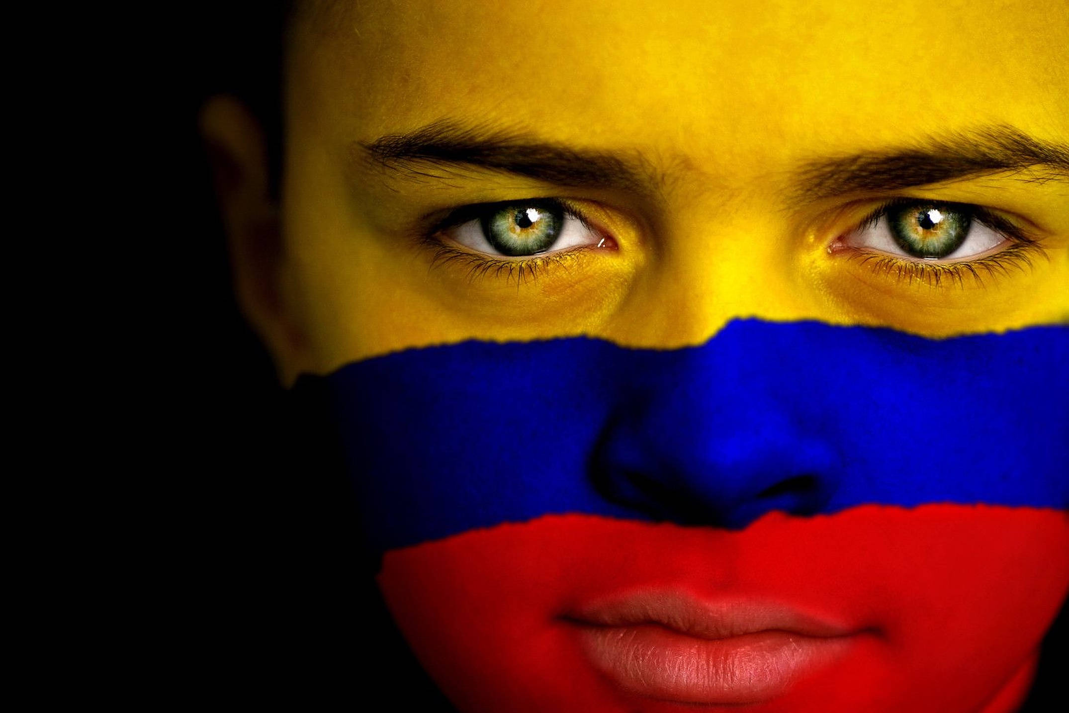 Colombia 桌布 壁紙 — 特寫肖像，一個人的臉上畫著哥倫比亞國旗，背景為黑色。
