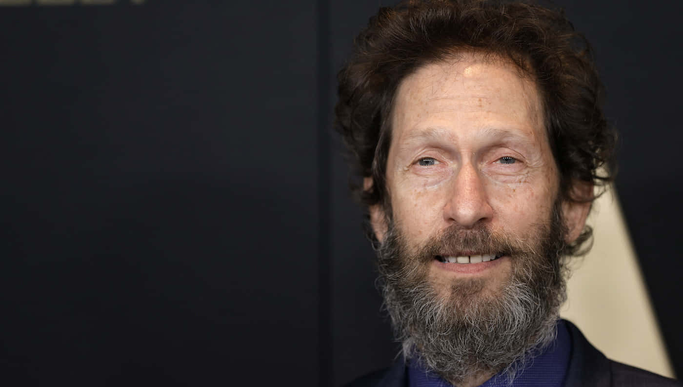 Tim Blake Nelson 桌布 壁紙 — 特寫肖像，一名留著棕色捲髮和灰白鬍鬚的男子在深色背景下溫暖微笑。
