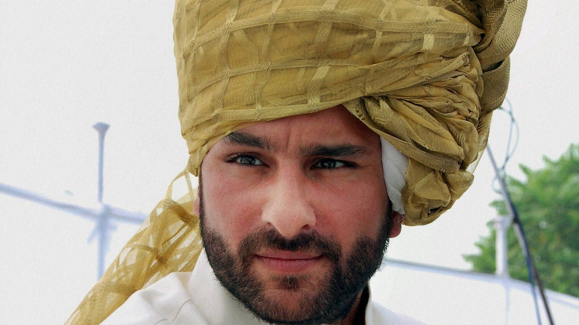 Saif Ali Khan 桌布 壁紙 — 特寫肖像，一名留鬍鬚的男子戴著質感金色頭巾，背景為明亮的戶外環境。