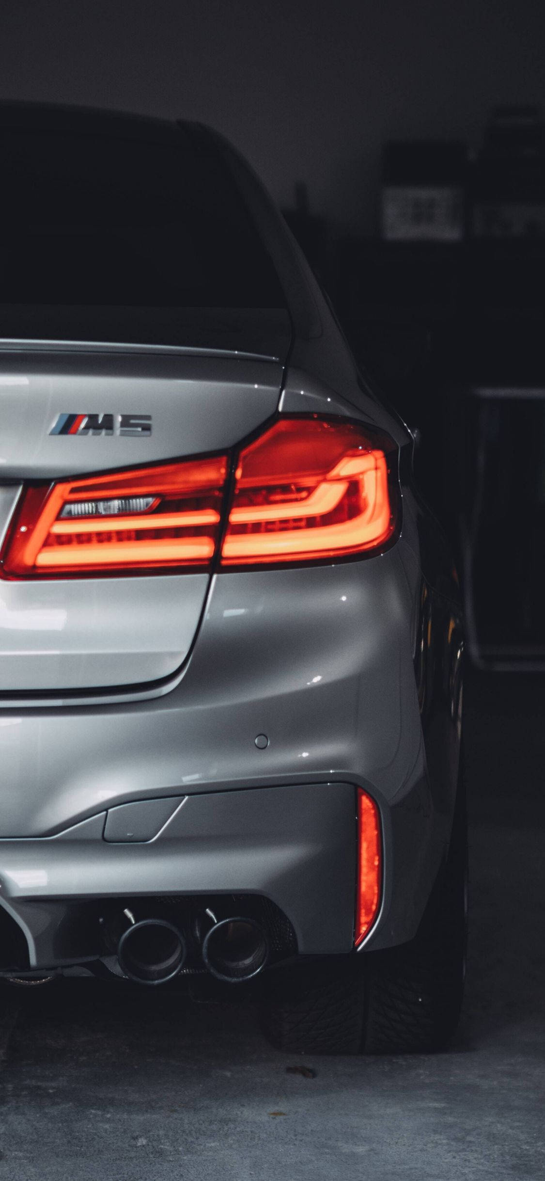 汽車 iPhone 壁紙 桌布 壁紙 — 特寫銀色BMW M5在昏暗車庫中發光的紅色尾燈和雙排氣管。