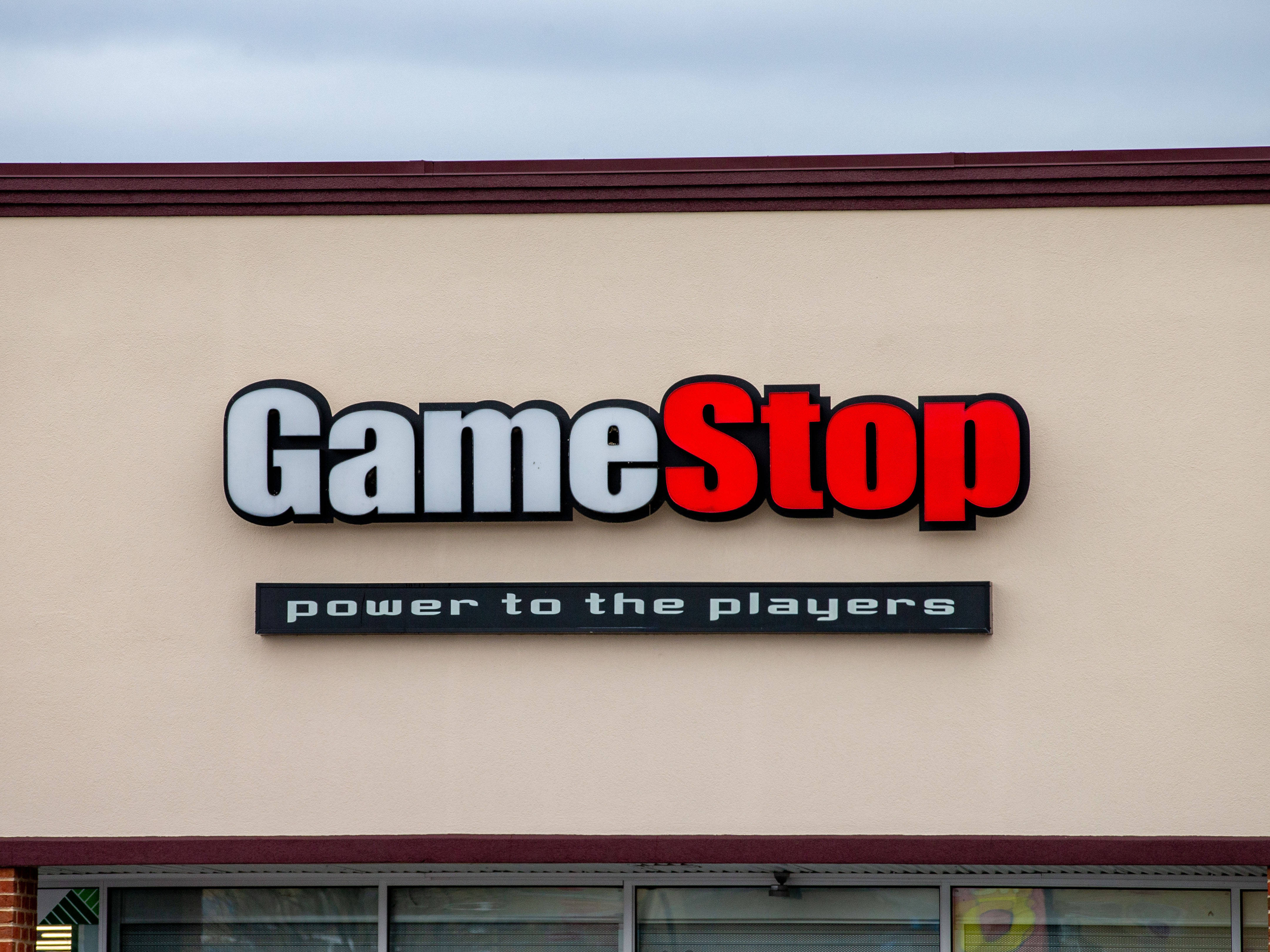 GameStop 桌布 壁紙 — 特寫鏡頭：GameStop 商店招牌，以粗體紅白字體呈現，背景為米色建築外牆，天空多雲。