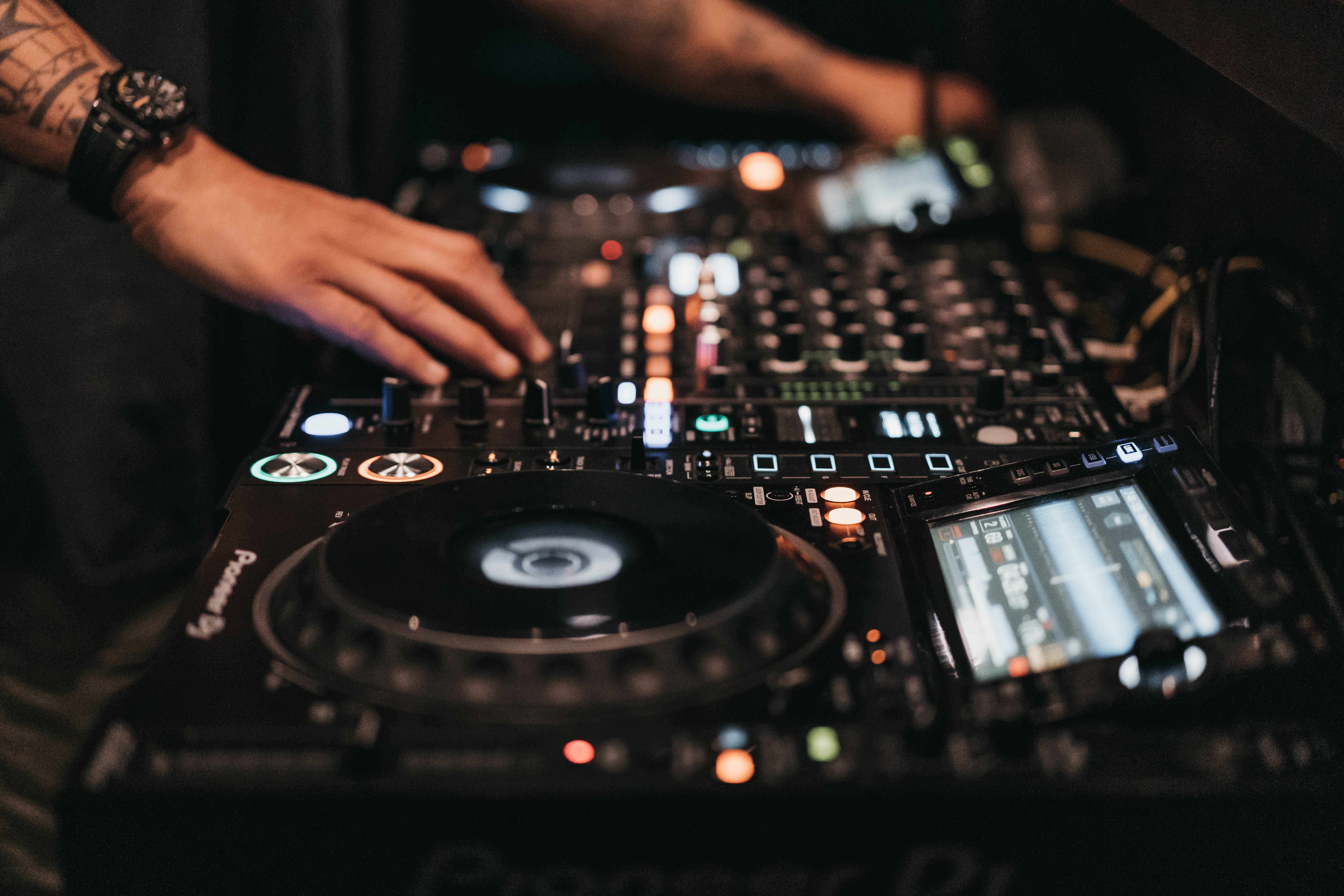 DJ 桌布 壁紙 — 特寫鏡頭：一位DJ刺青的手在昏暗的俱樂部中調整發光的黑色Pioneer DJ控制器上的旋鈕。