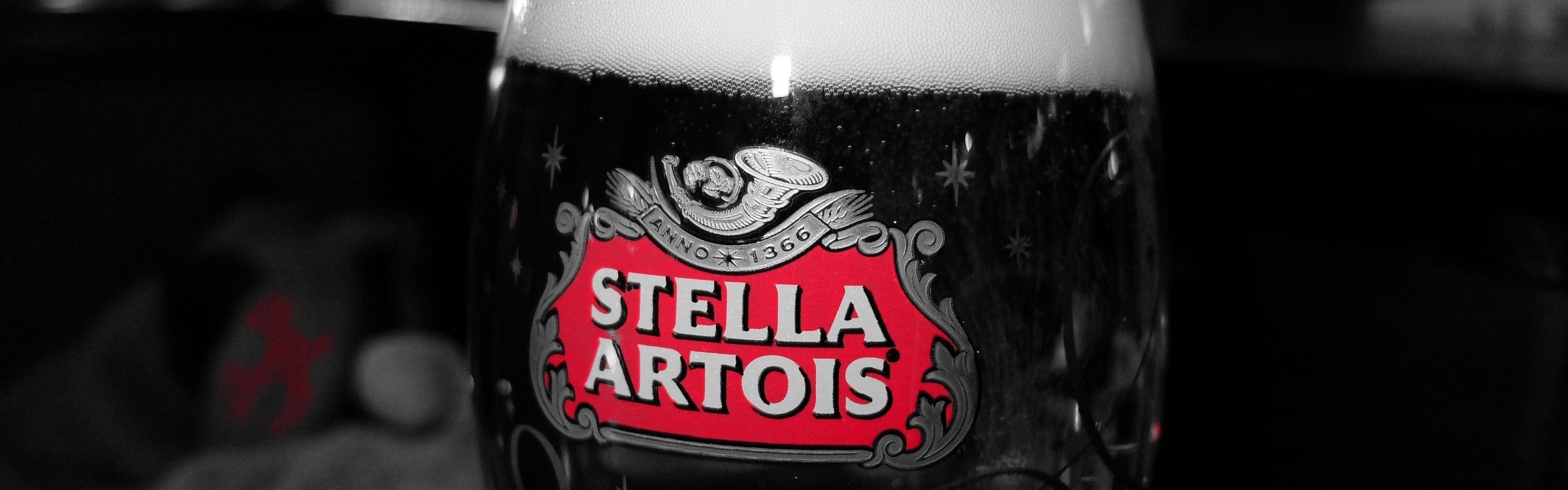 飲料 桌布 壁紙 — 特寫鏡頭：一個Stella Artois啤酒杯，帶有泡沫豐富的頂部和華麗的標誌，在黑暗的單色背景下發出紅光。