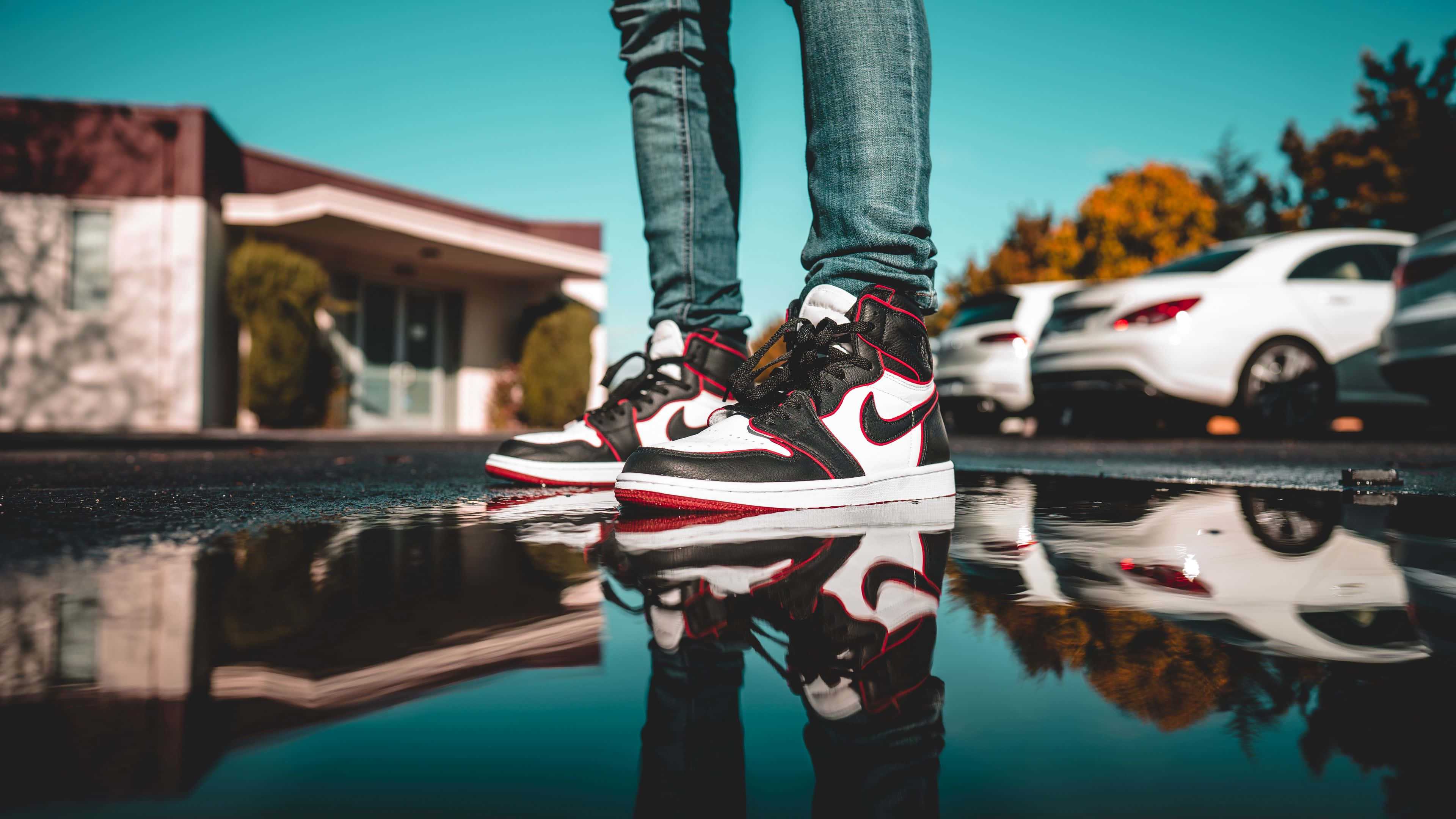 Sneaker 桌布 壁紙 — 特寫鏡頭：一個人的腳穿著黑色、紅色和白色的運動鞋，在陽光明媚的日子裡倒映在水坑中，背景有汽車和房屋。