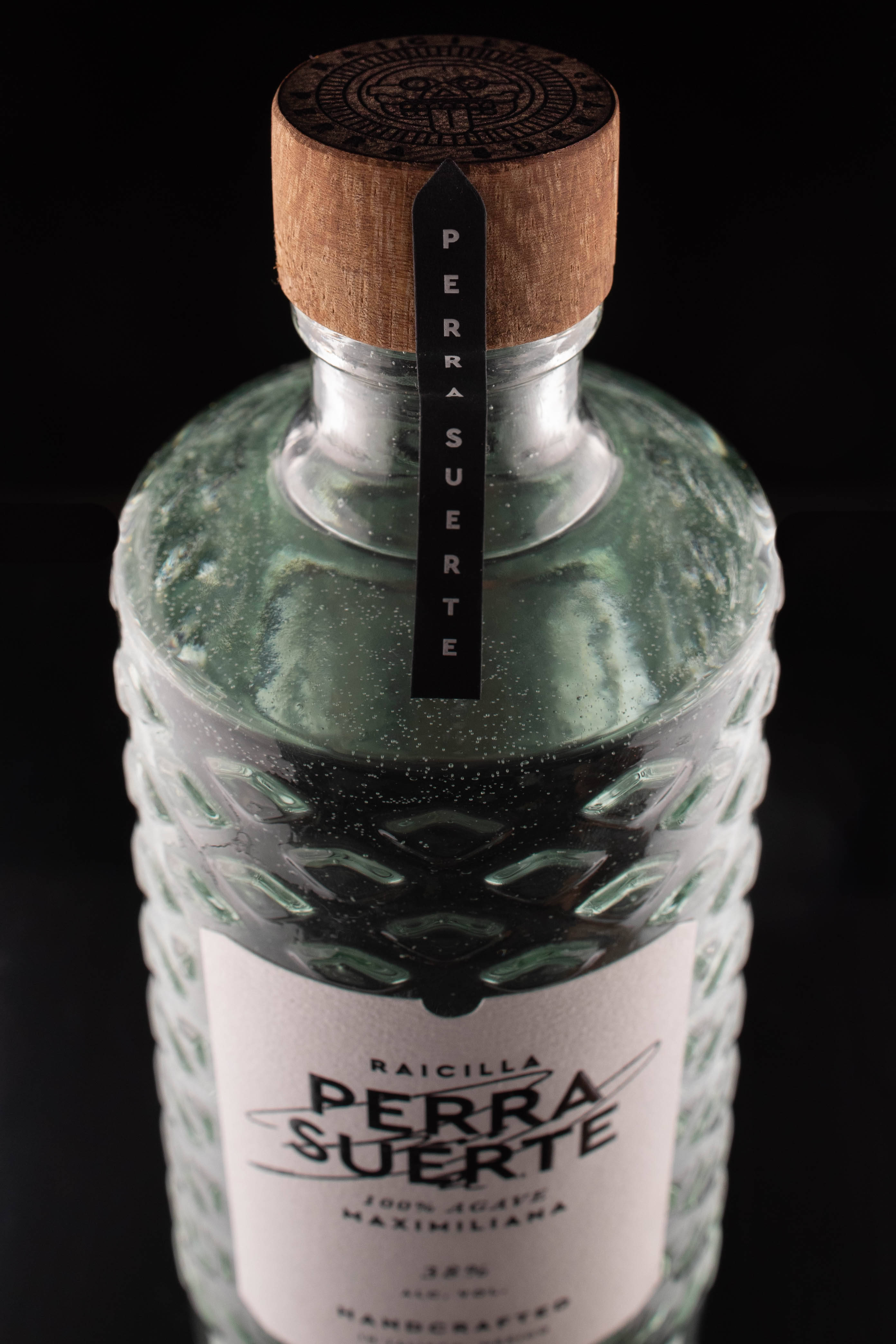 alcohol 桌布 壁紙 — 特寫鏡頭：一個帶有木蓋的紋理綠色玻璃瓶Raicilla Perra Suerte，置於黑色背景前。