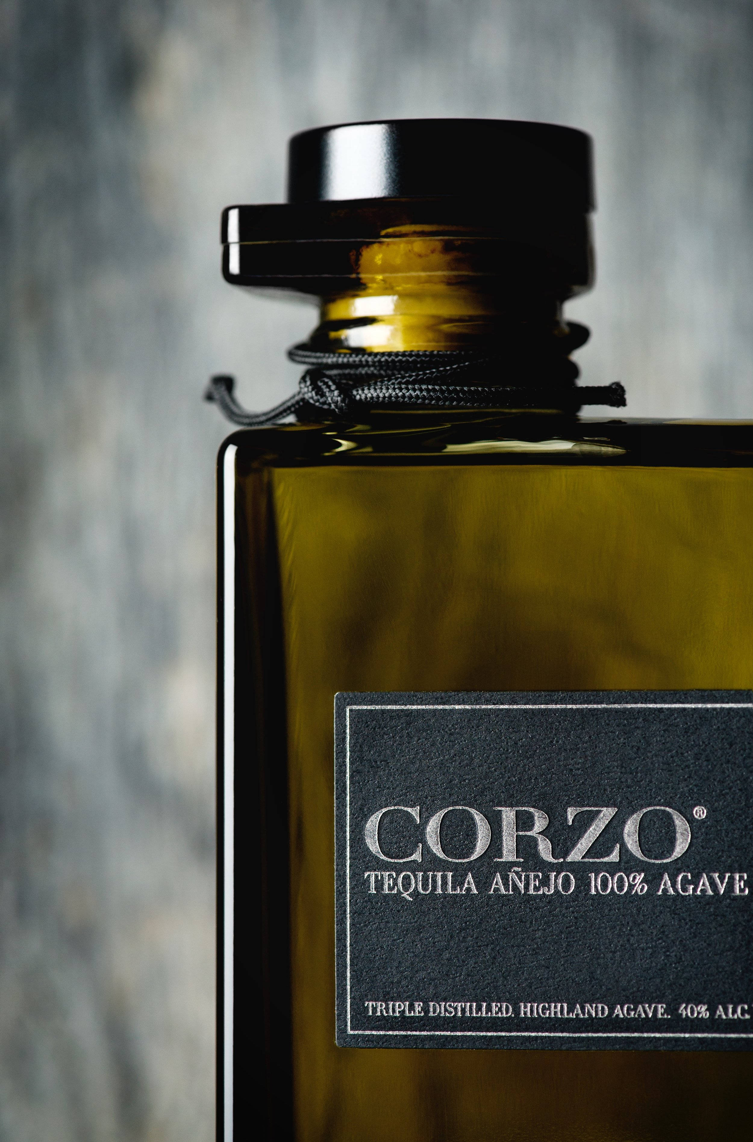 Tequila 桌布 壁紙 — 特寫鏡頭：一個時尚的琥珀色龍舌蘭酒瓶，帶有黑色瓶蓋和標籤，標籤上寫著「Corzo Tequila Añejo 100% Agave」，背景為紋理灰色。