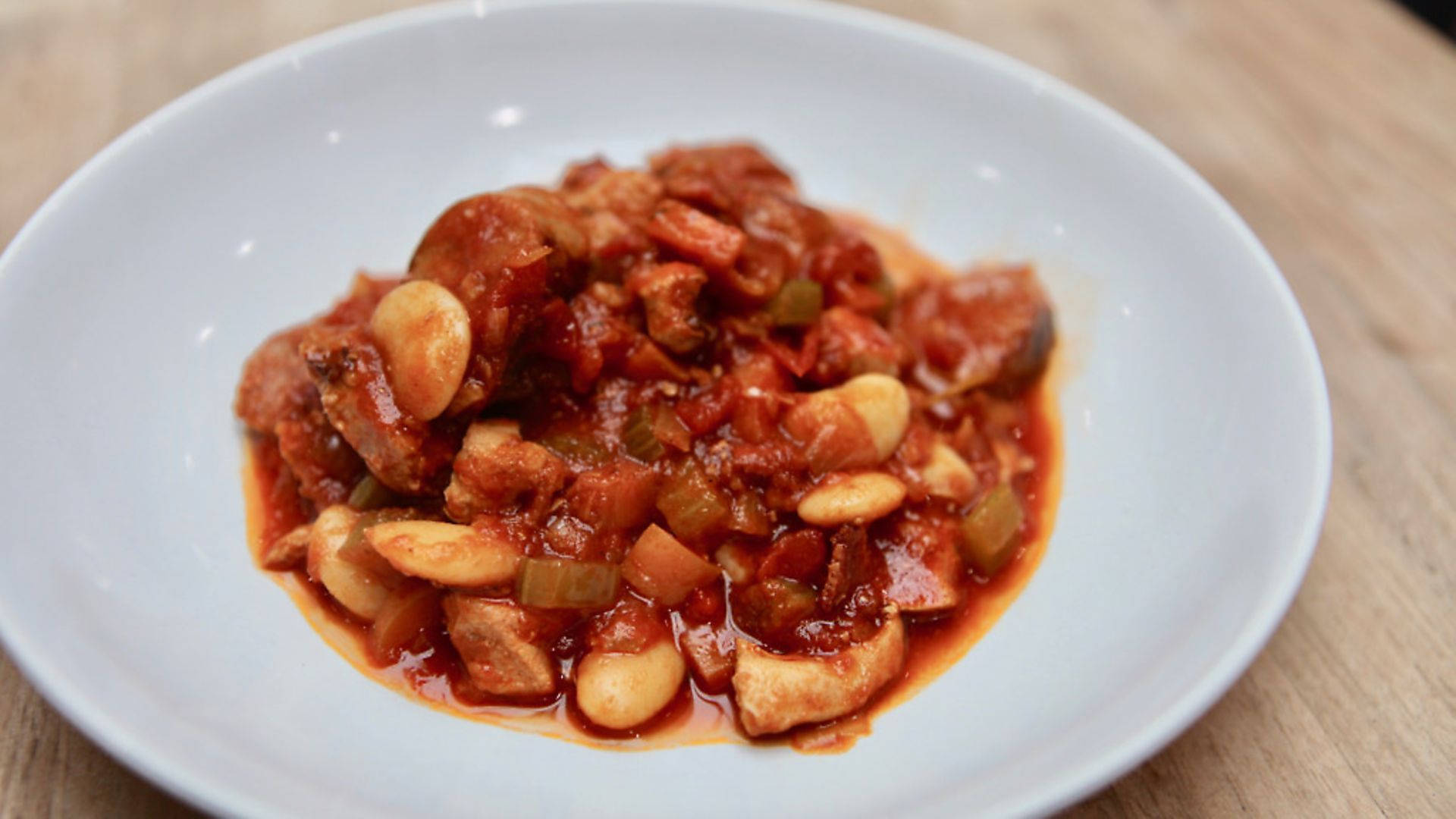 Delicious Food 桌布 壁紙 — 特寫鏡頭：一個白色碗中的豐盛白豆燉菜，配有香腸和蔬菜塊，放在木桌上。