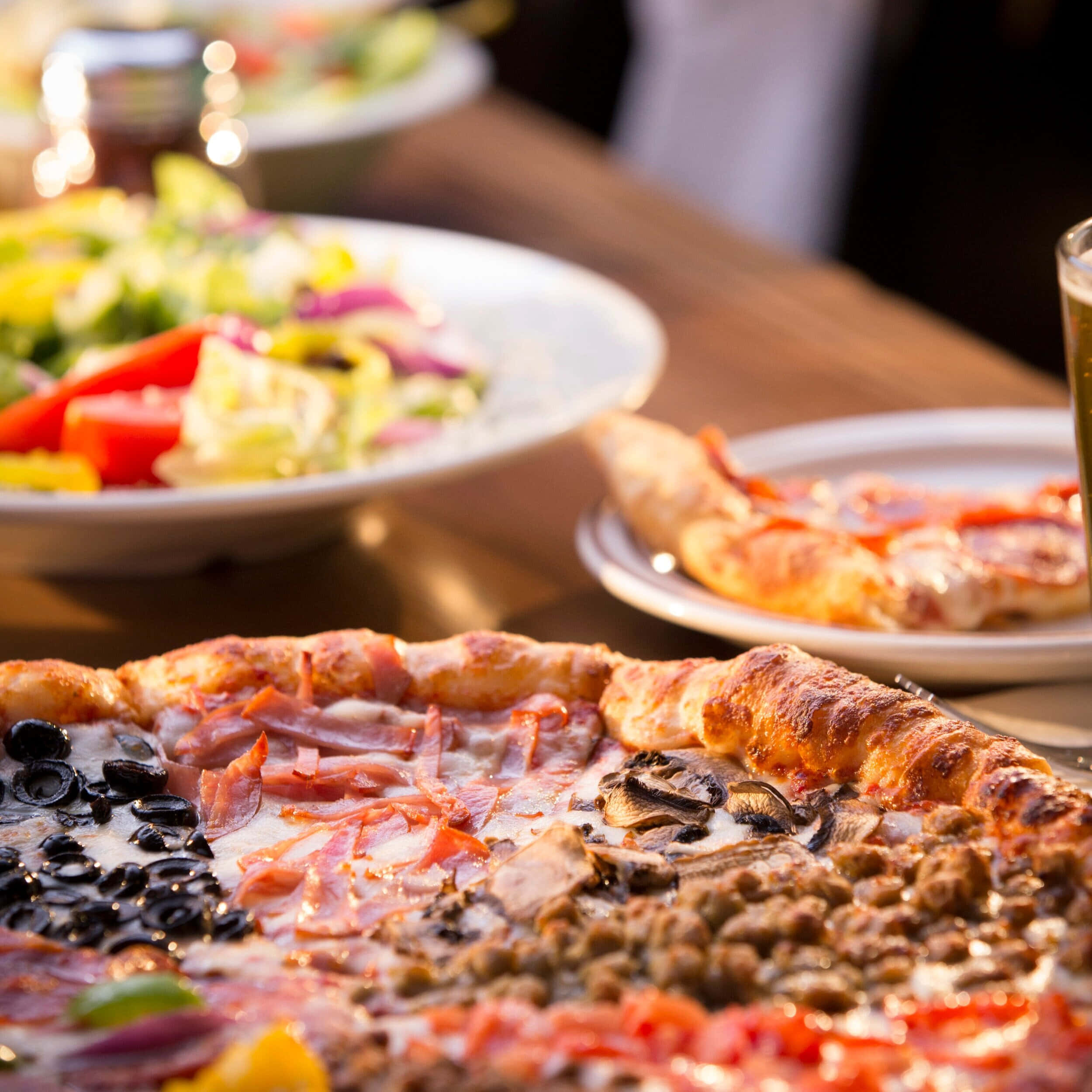 Pizza 桌布 壁紙 — 特寫鏡頭：一個美味滿載的披薩，上面有橄欖、火腿、蘑菇和香腸，鋪上融化起司，放在木桌上，背景有沙拉和飲料。