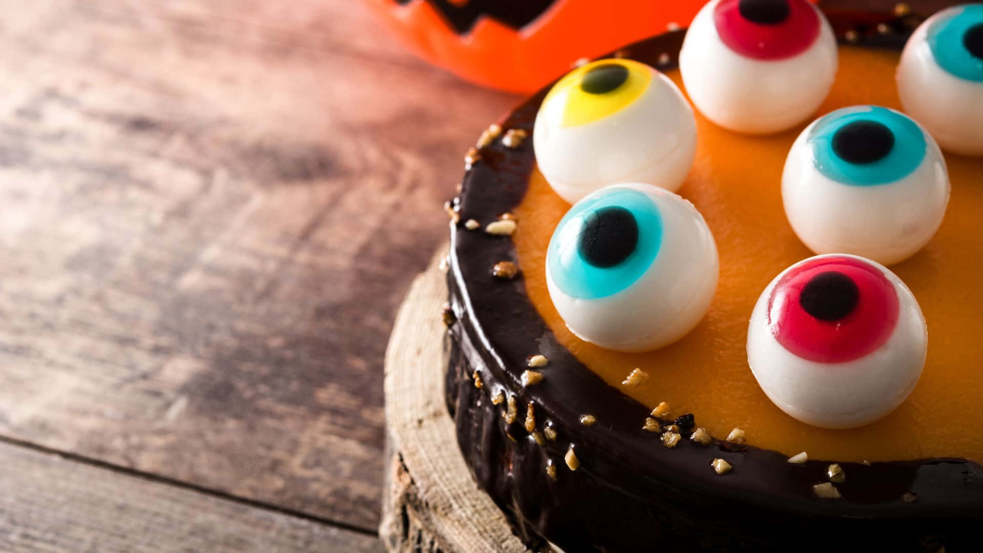 Halloween Cake 桌布 壁紙 — 特寫鏡頭：一個萬聖節蛋糕，上面鋪滿色彩繽紛的糖果眼睛和巧克力甘納許，放在質樸的木板上。