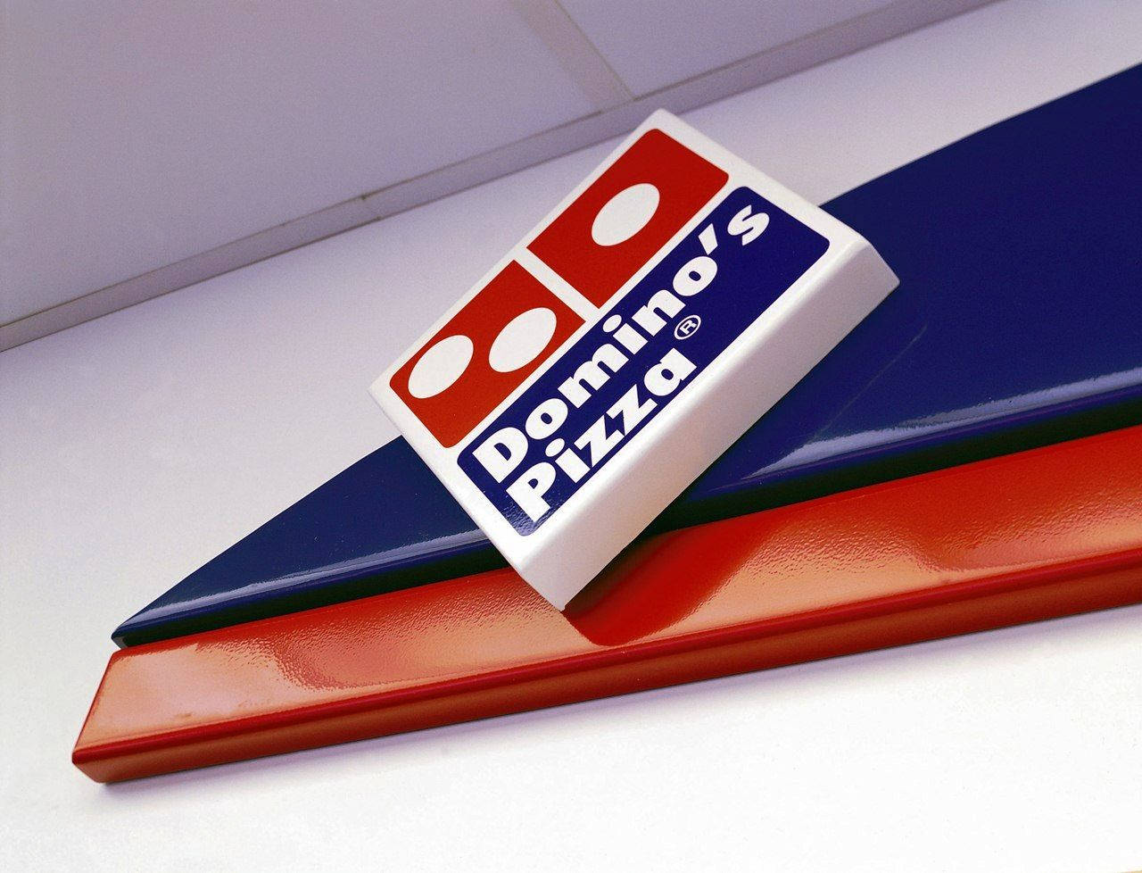 Dominos Pizza 桌布 壁紙 — 特寫鏡頭：一塊 Domino's Pizza 標誌牌擱在紅色和藍色傾斜面板上，背景為白色天花板。