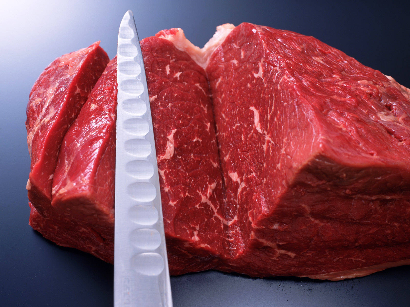 meat 桌布 壁紙 — 特寫鏡頭：一塊生牛肉切塊，旁邊放著一把鋸齒刀，置於深色表面上。