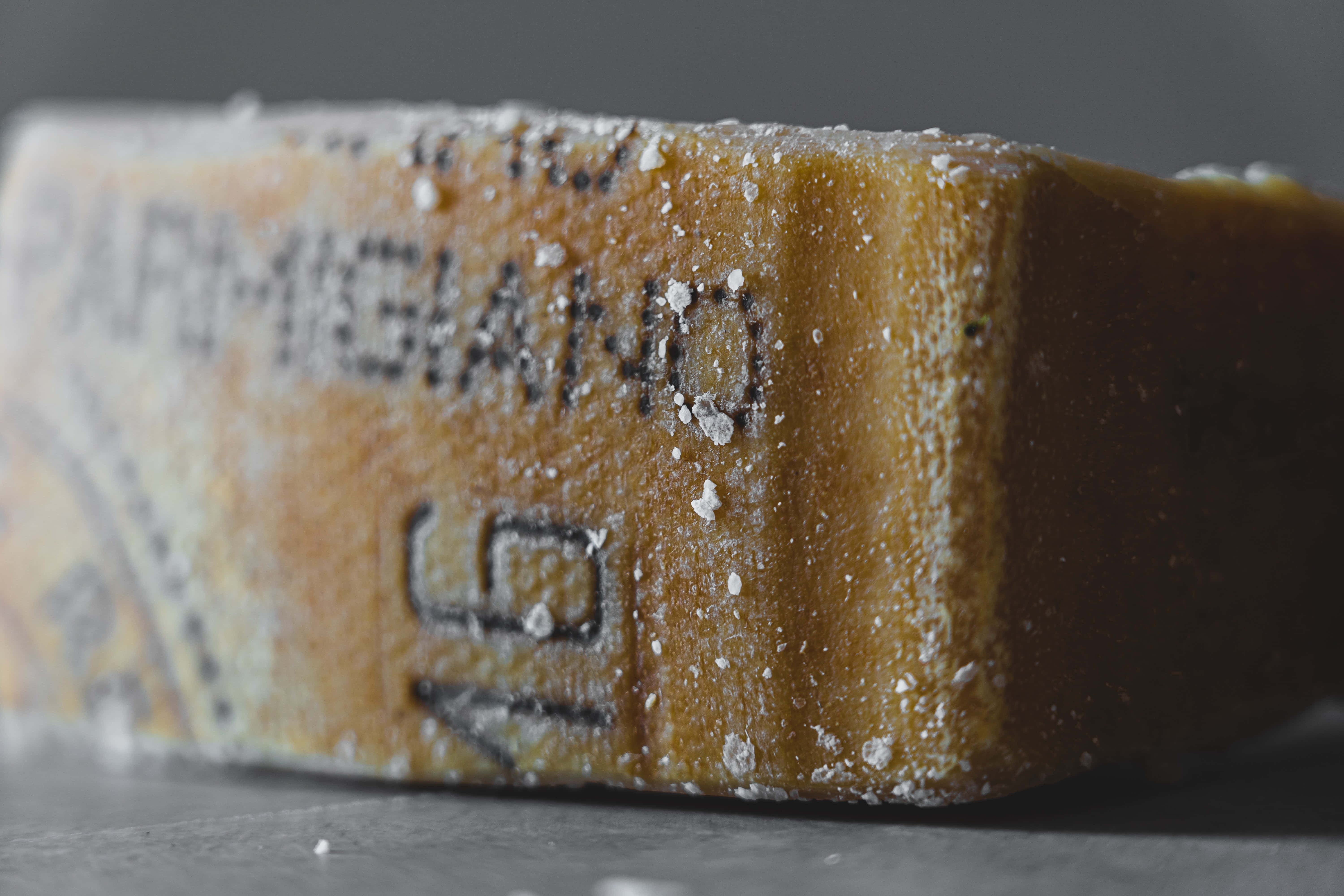 Cheese 桌布 壁紙 — 特寫鏡頭：一塊鄉村風格的帕瑪森雷吉亞諾起司楔形塊，撒上細白晶體，置於深灰色背景上。