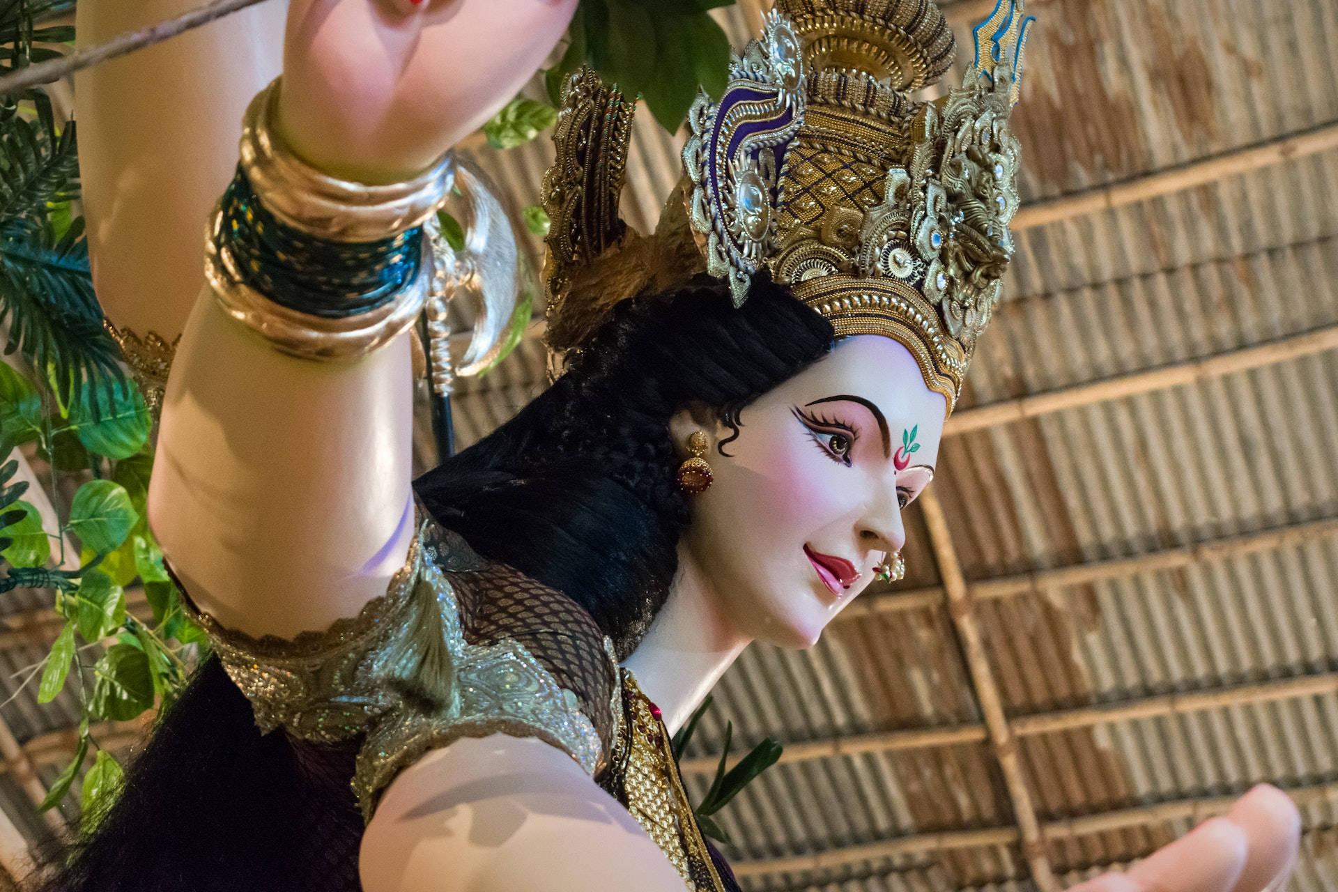 Durga 桌布 壁紙 — 特寫鏡頭，一座裝飾華麗的女神雕像，戴著金色頭飾和珠寶，背景是茅草屋頂和綠植。