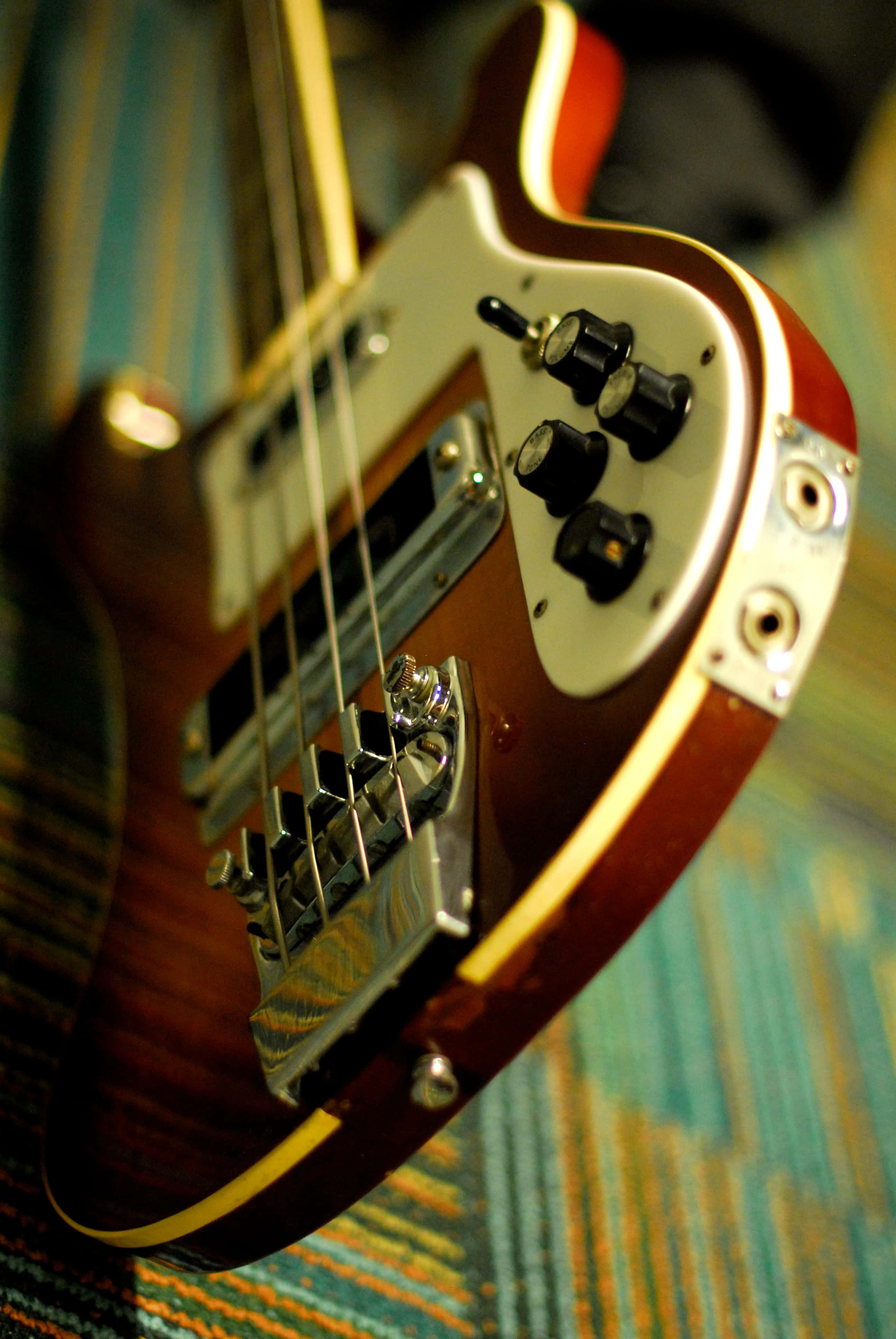 Bass Guitar 桌布 壁紙 — 特寫鏡頭：一把帶有鍍鉻硬件的旭日色低音吉他，擱在色彩繽紛的條紋地毯上。