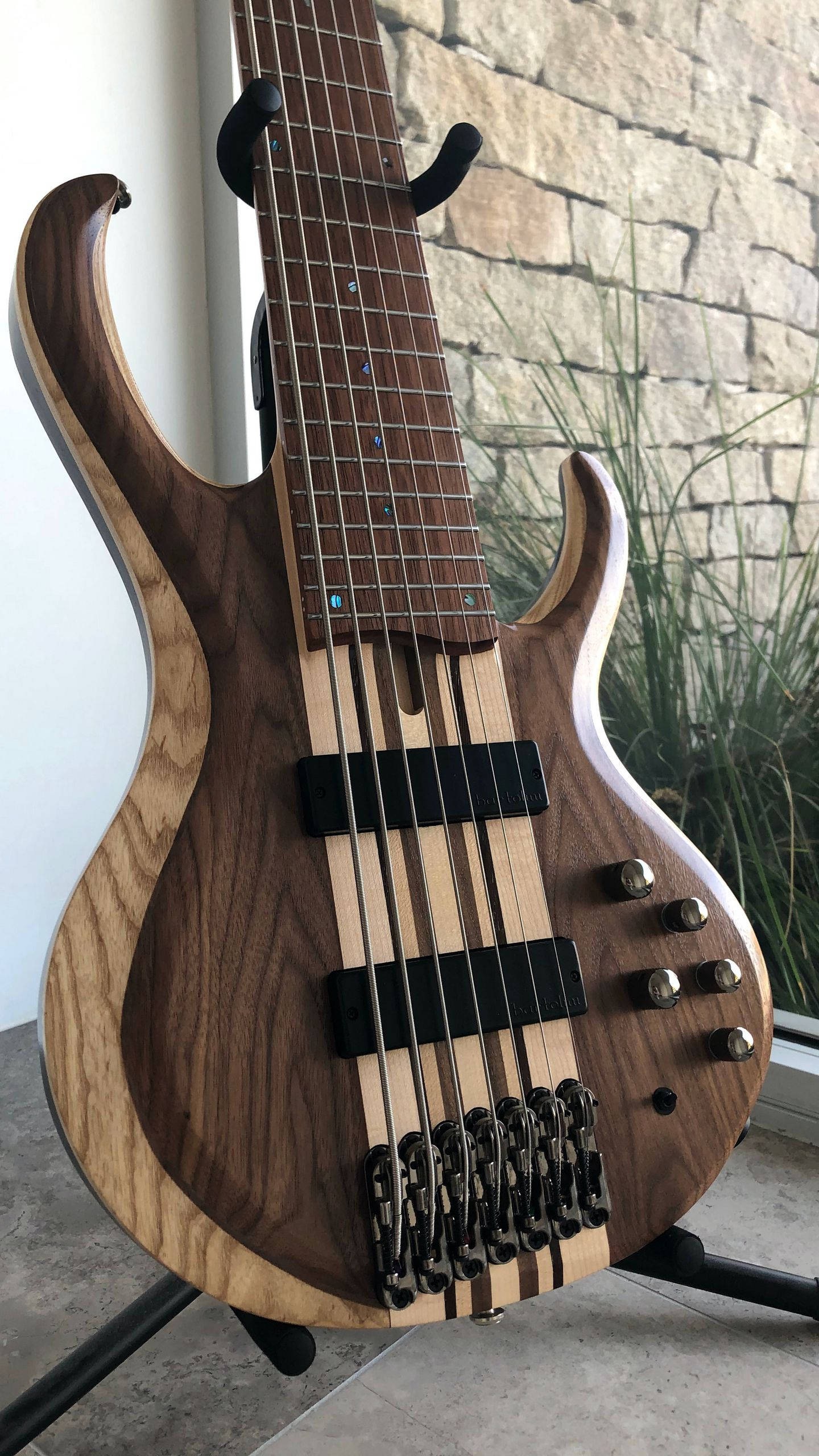 Brown iPhone 桌布 壁紙 — 特寫鏡頭：一把訂製五弦低音吉他，擁有豐富木紋和黑色硬體，擱在支架上，背景是石牆和綠植。