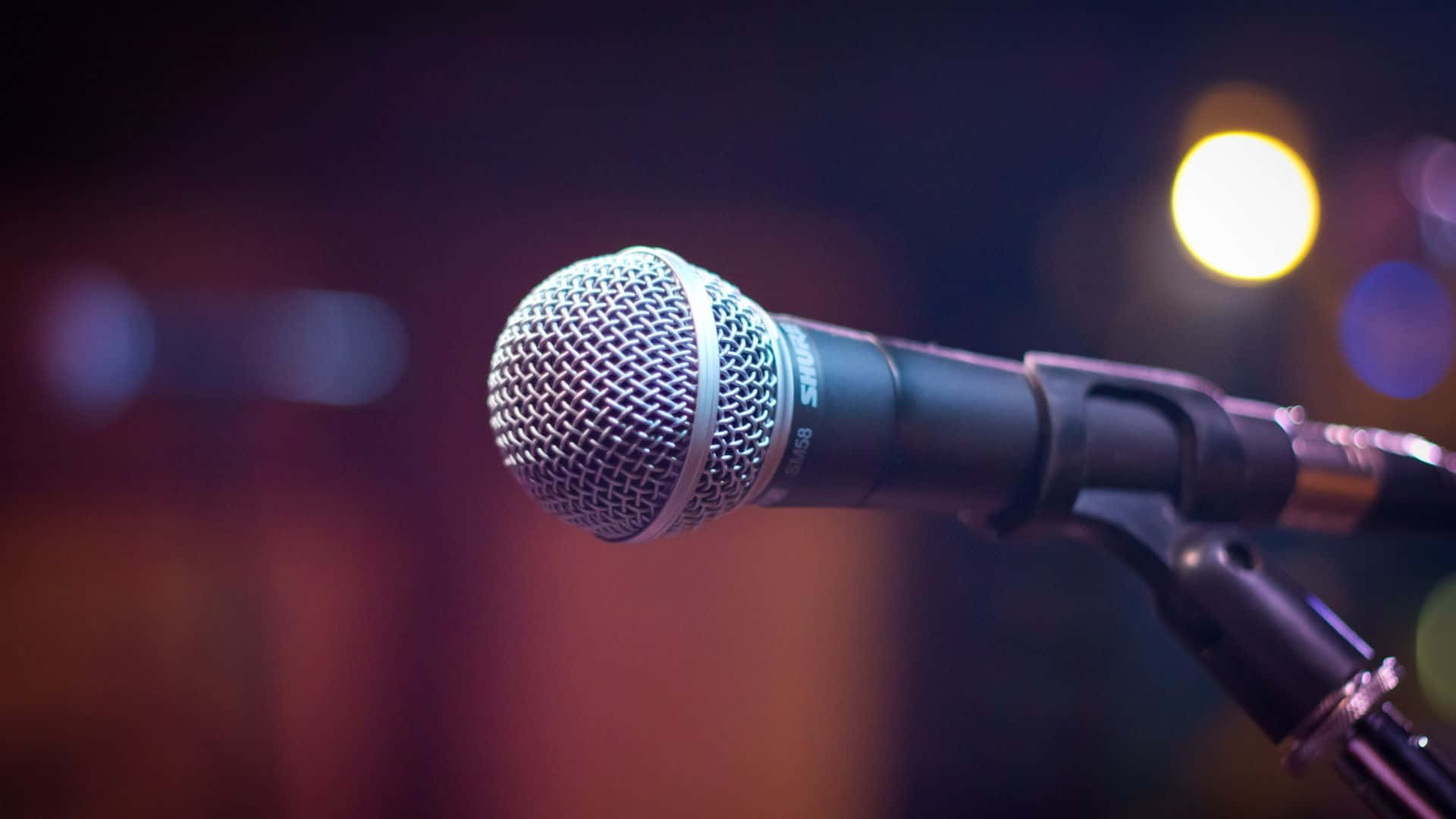Microphone 桌布 壁紙 — 特寫鏡頭：一支Shure麥克風放在麥克風架上，處於溫暖舞台燈光下，背景模糊。