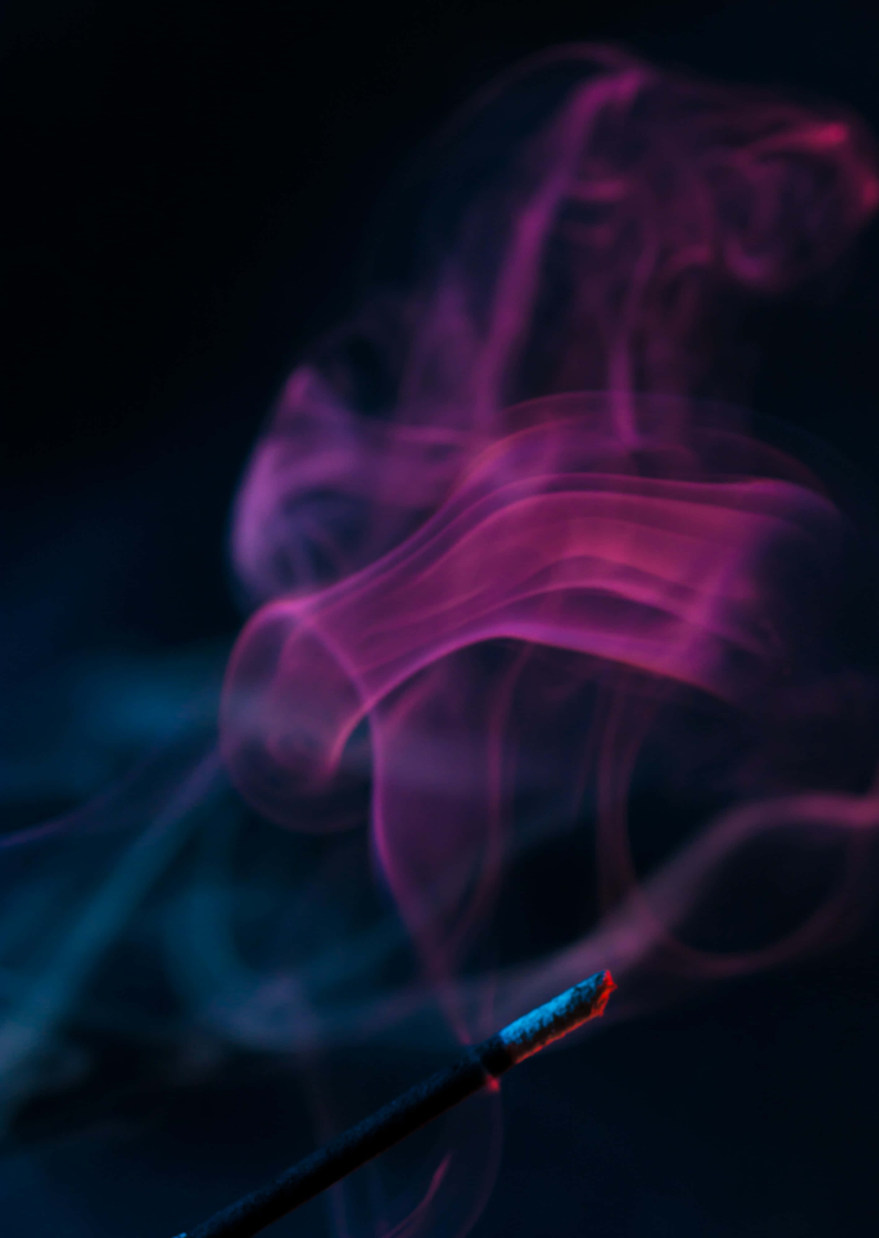 Pink Smoke 桌布 壁紙 — 特寫鏡頭，一支發光的香在黑暗背景下釋放出旋轉的紫色和藍色煙霧。