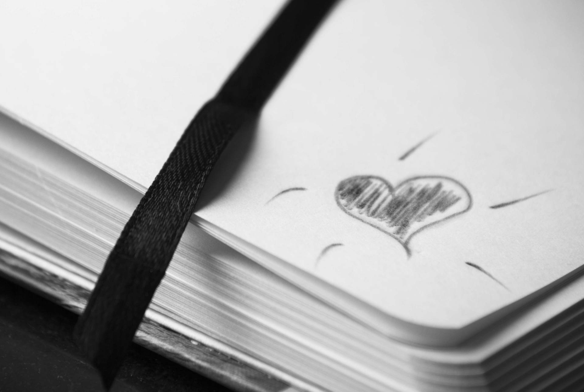 Heart 桌布 壁紙 — 特寫鏡頭：一本帶有黑色彈性帶的素描本，空白頁上有一個簡單的鉛筆繪製的心形，散發出線條。