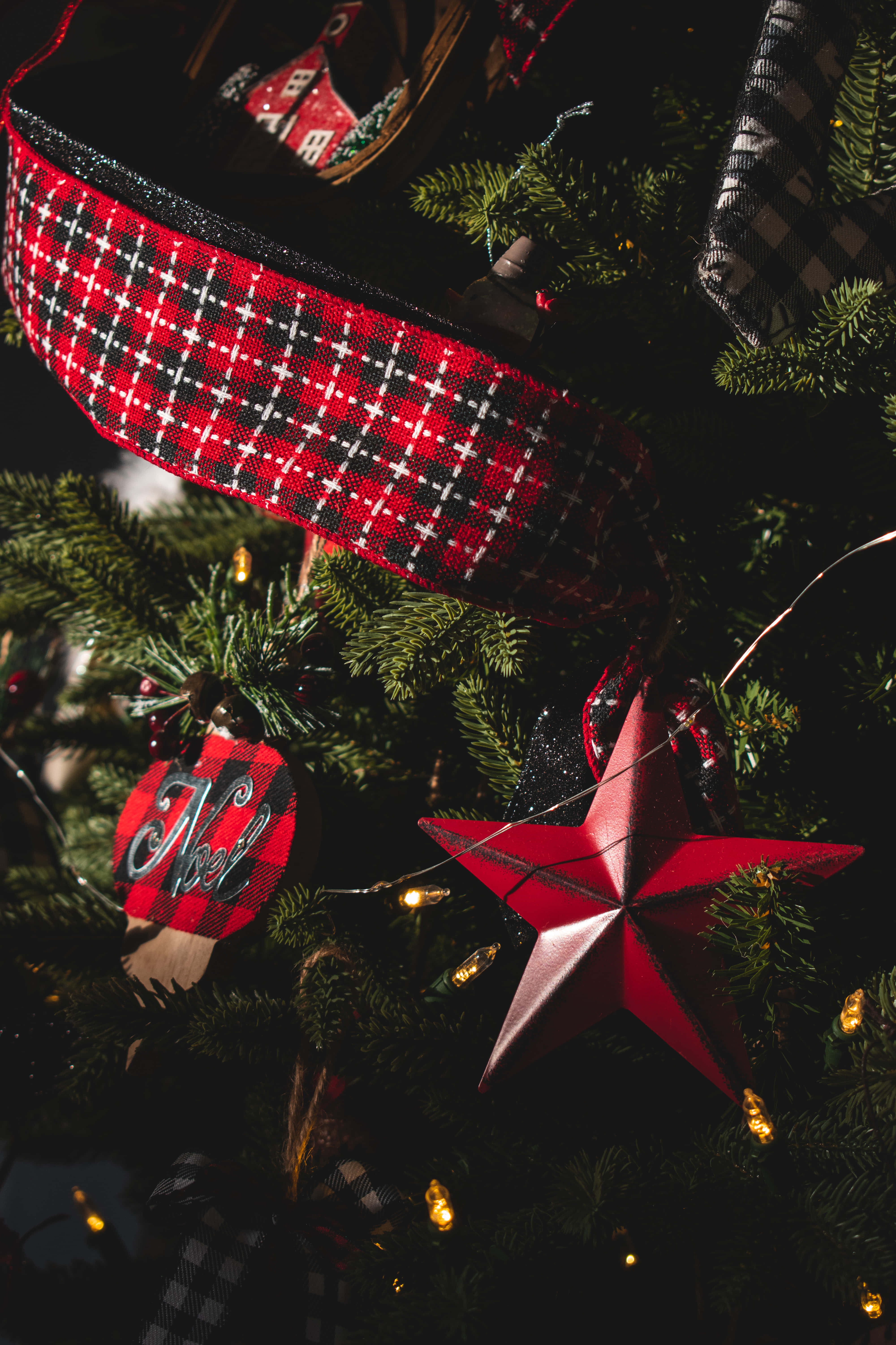 Christmas Tree iPhone 桌布 壁紙 — 特寫鏡頭：一棵聖誕樹，裝飾著紅色格子絲帶、紅色星星飾物和溫暖的仙女燈，背景是深綠色松針。