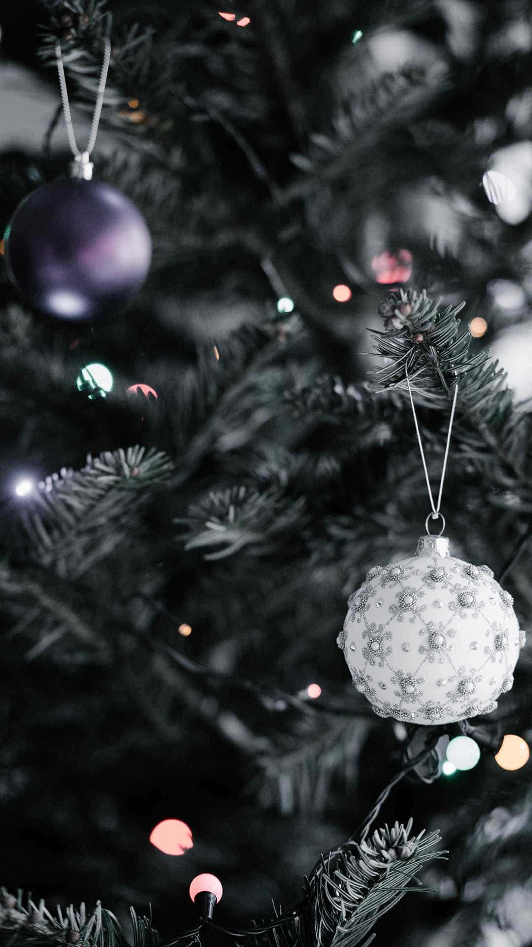 Christmas Tree iPhone 桌布 壁紙 — 特寫鏡頭：一棵裝飾好的聖誕樹，帶有白色珠飾和紫色聖誕球，背景是模糊的節日燈光。