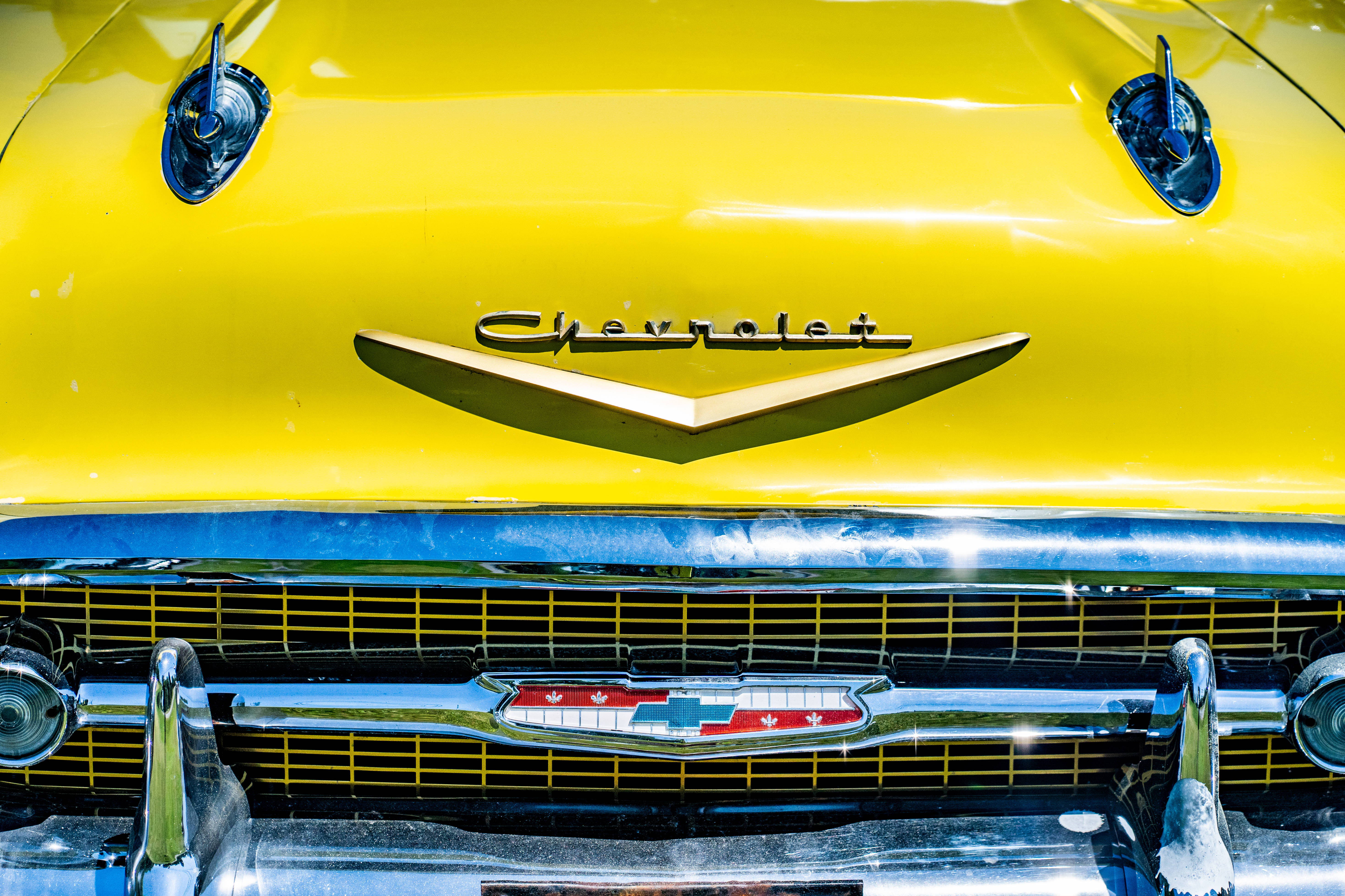 Chevrolet Logo 桌布 壁紙 — 特寫鏡頭：一輛復古黃色雪佛蘭汽車在明亮陽光下閃閃發光的引擎蓋標誌和鍍鉻格柵。