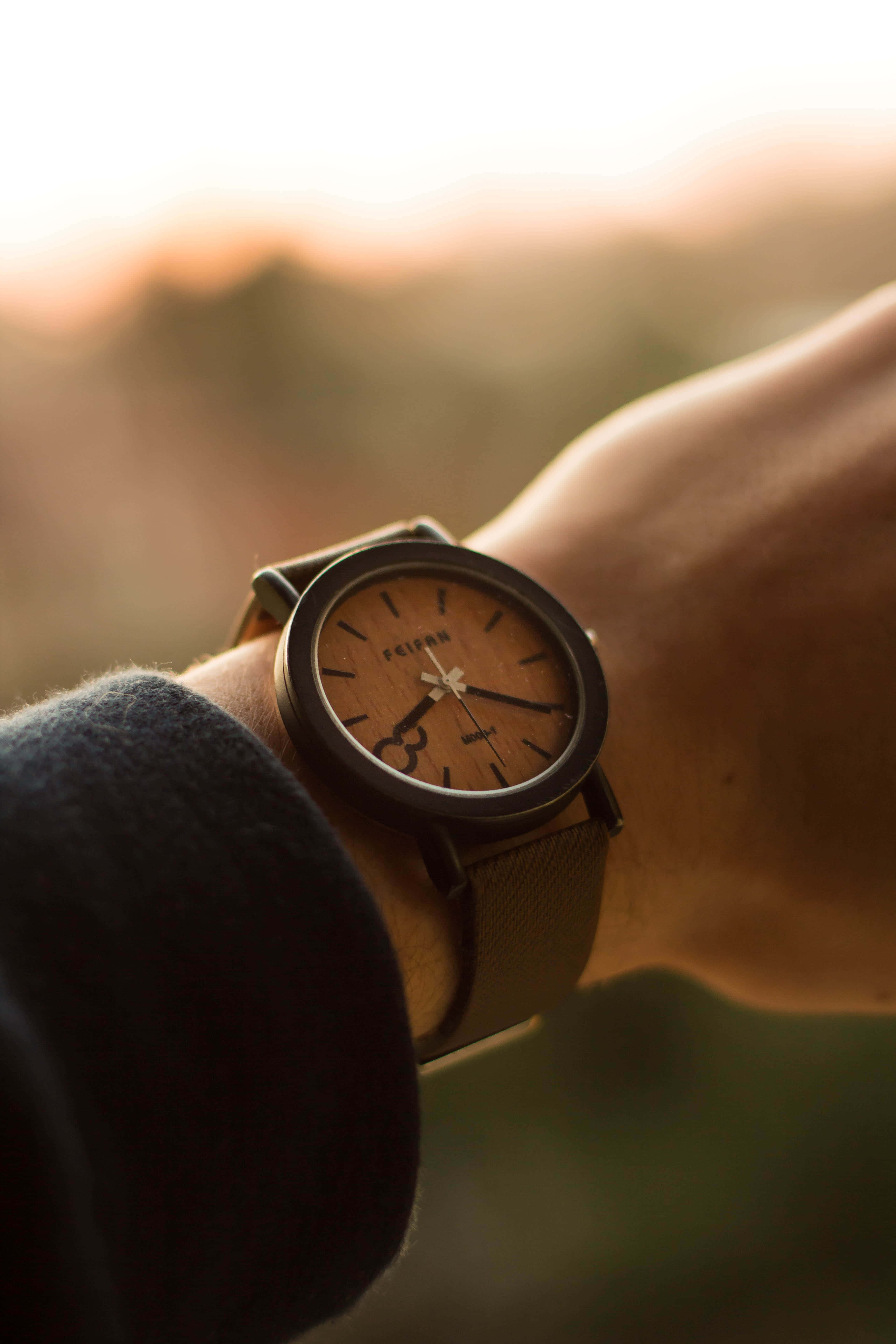 Clock 桌布 壁紙 — 特寫鏡頭：一隻木製手錶戴在人的手腕上，背景是溫暖的日落。