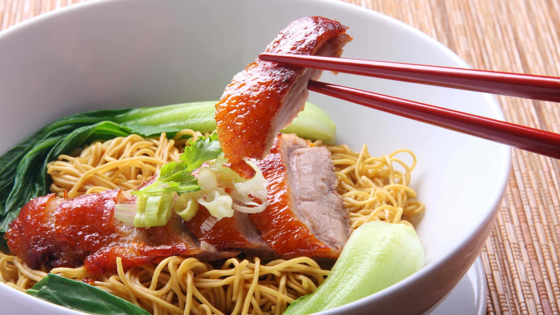 Food Photography 桌布 壁紙 — 特寫鏡頭，一隻油亮的烤鴨被紅色筷子從一碗黃色麵條和綠色青江菜中夾起。