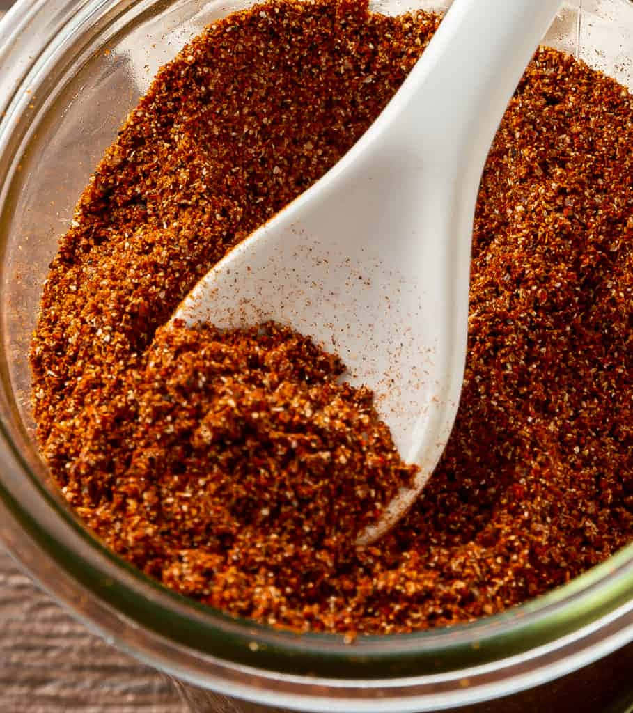 Red Chili Powder 桌布 壁紙 — 特寫鏡頭，一隻白色湯匙從透明玻璃罐中舀出濃郁的紅棕色香料混合物。