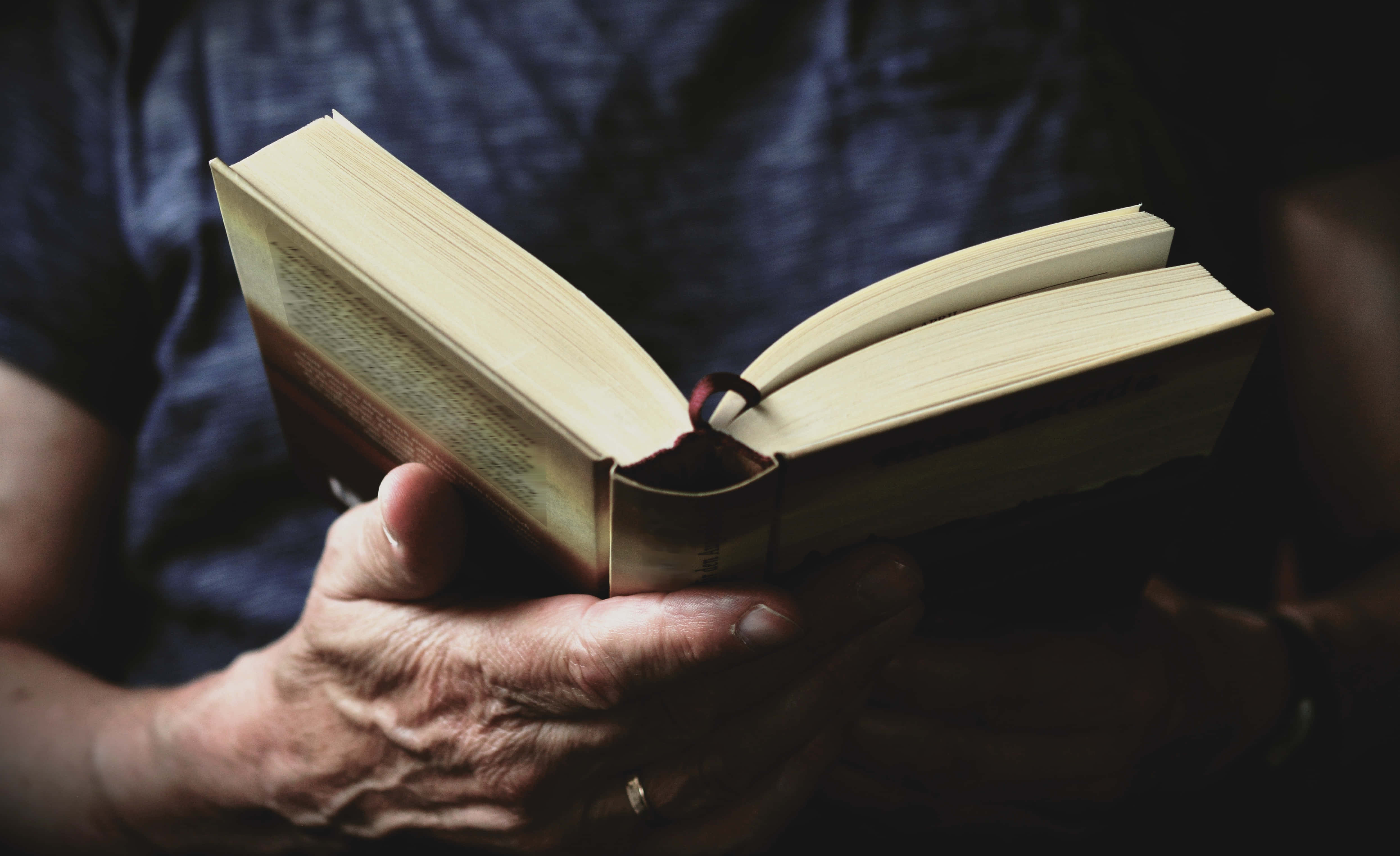 Reading 桌布 壁紙 — 特寫鏡頭，一雙歷經風霜的手在柔和暖光下捧著一本泛黃書頁的打開書本。