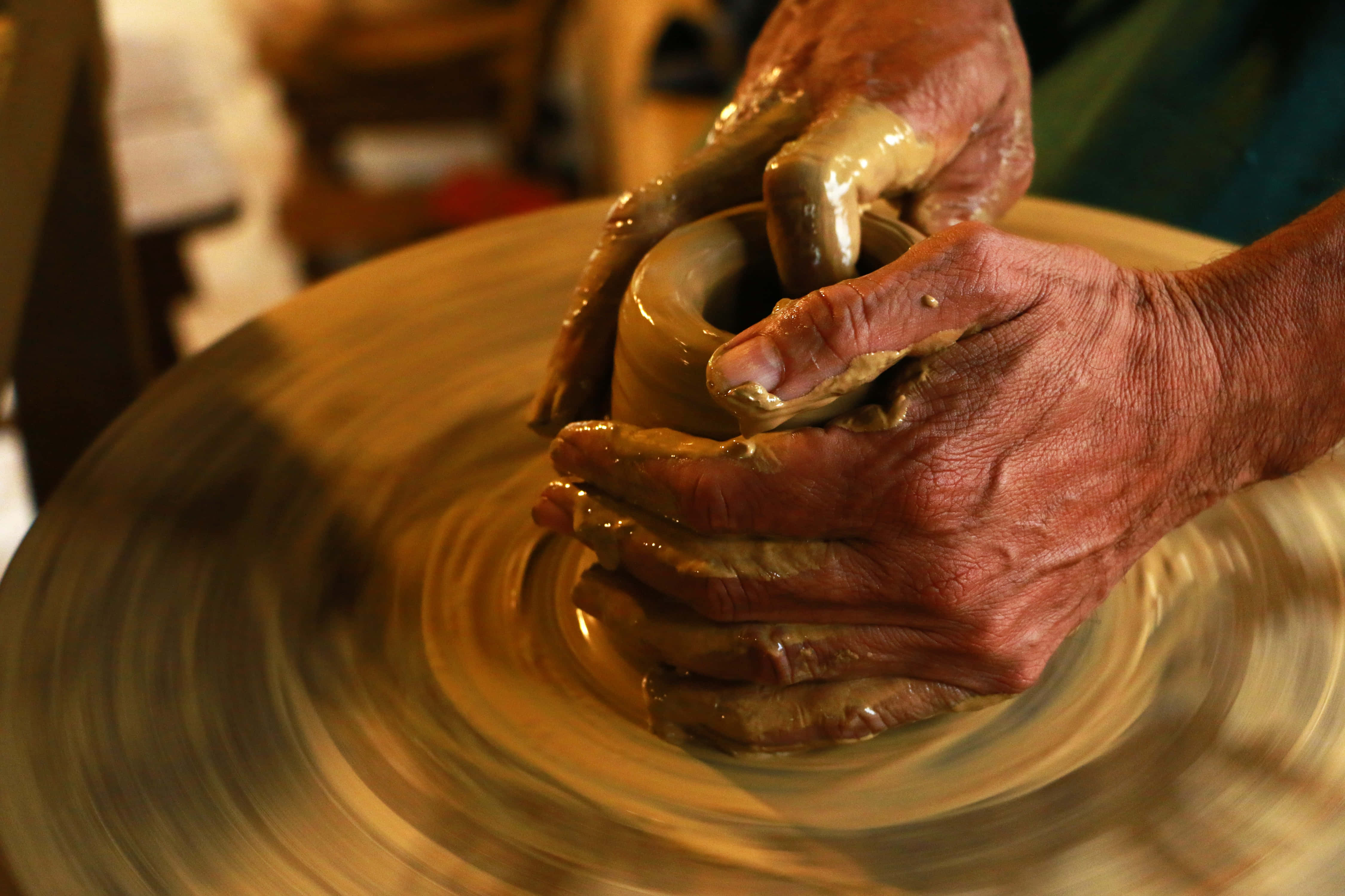 hands 桌布 壁紙 — 特寫鏡頭，一雙歷經風霜的手在溫暖的金色光線下，熟練地在旋轉的陶輪上塑造濕黏土。