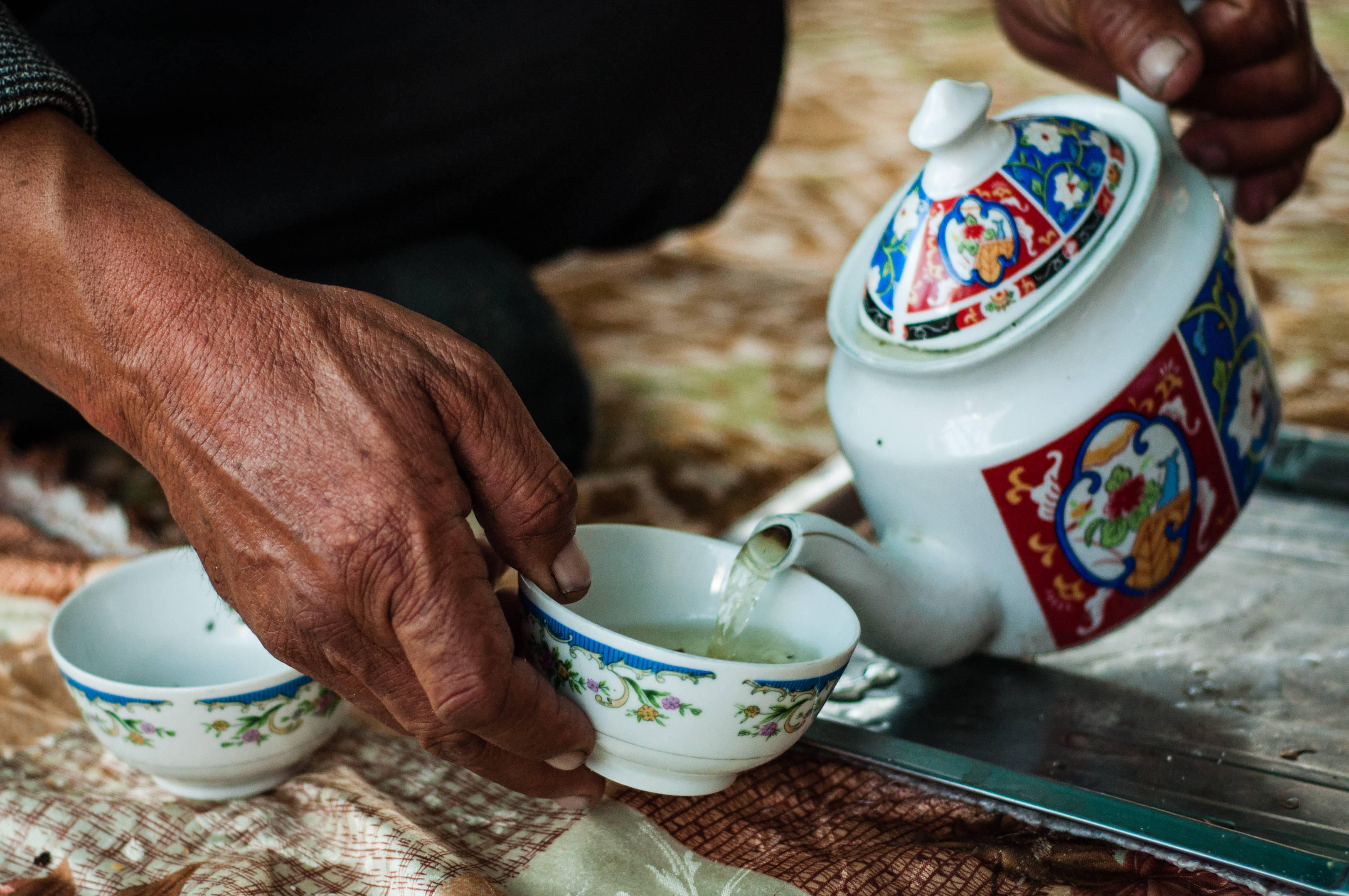 Tajikistan 桌布 壁紙 — 特寫鏡頭：一雙飽經風霜的手，將清澈的茶從色彩繽紛的陶瓷茶壺倒入相配的茶杯中，茶杯放在一塊有圖案的布上。