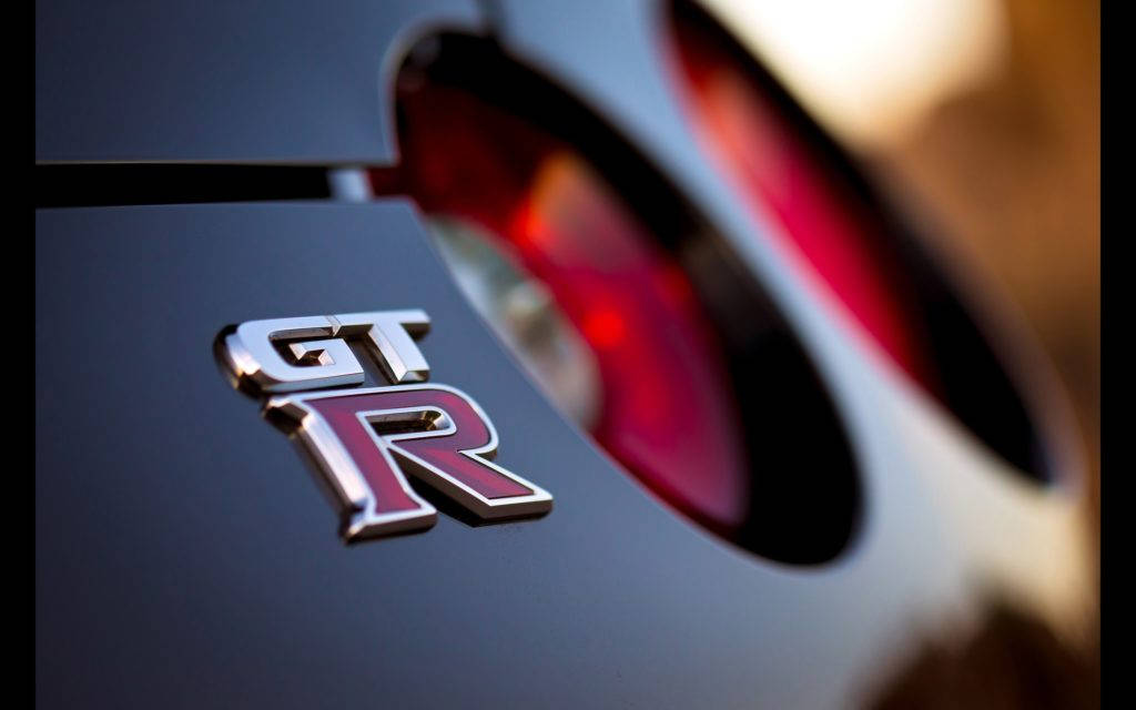 R 桌布 壁紙 — 特寫鏡頭：在溫暖逆光下，光滑黑色車身上的閃亮GT-R標誌，紅色尾燈模糊。