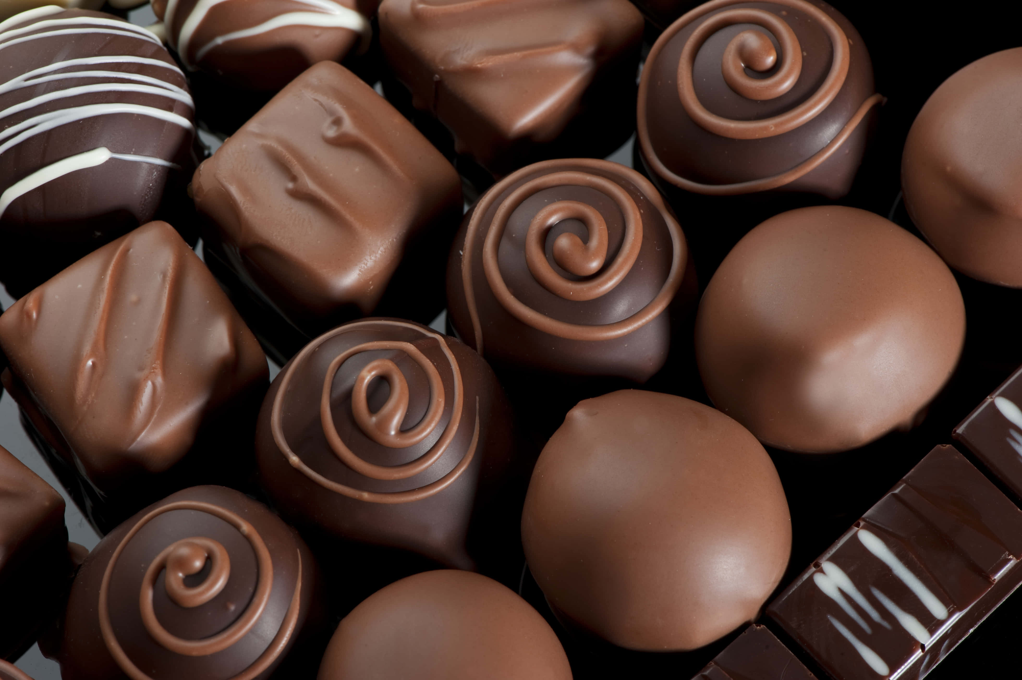 Brown Chocolate 桌布 壁紙 — 特寫鏡頭，展示各種光澤巧克力糖果，帶有漩渦和滴落效果，置於深色表面上。
