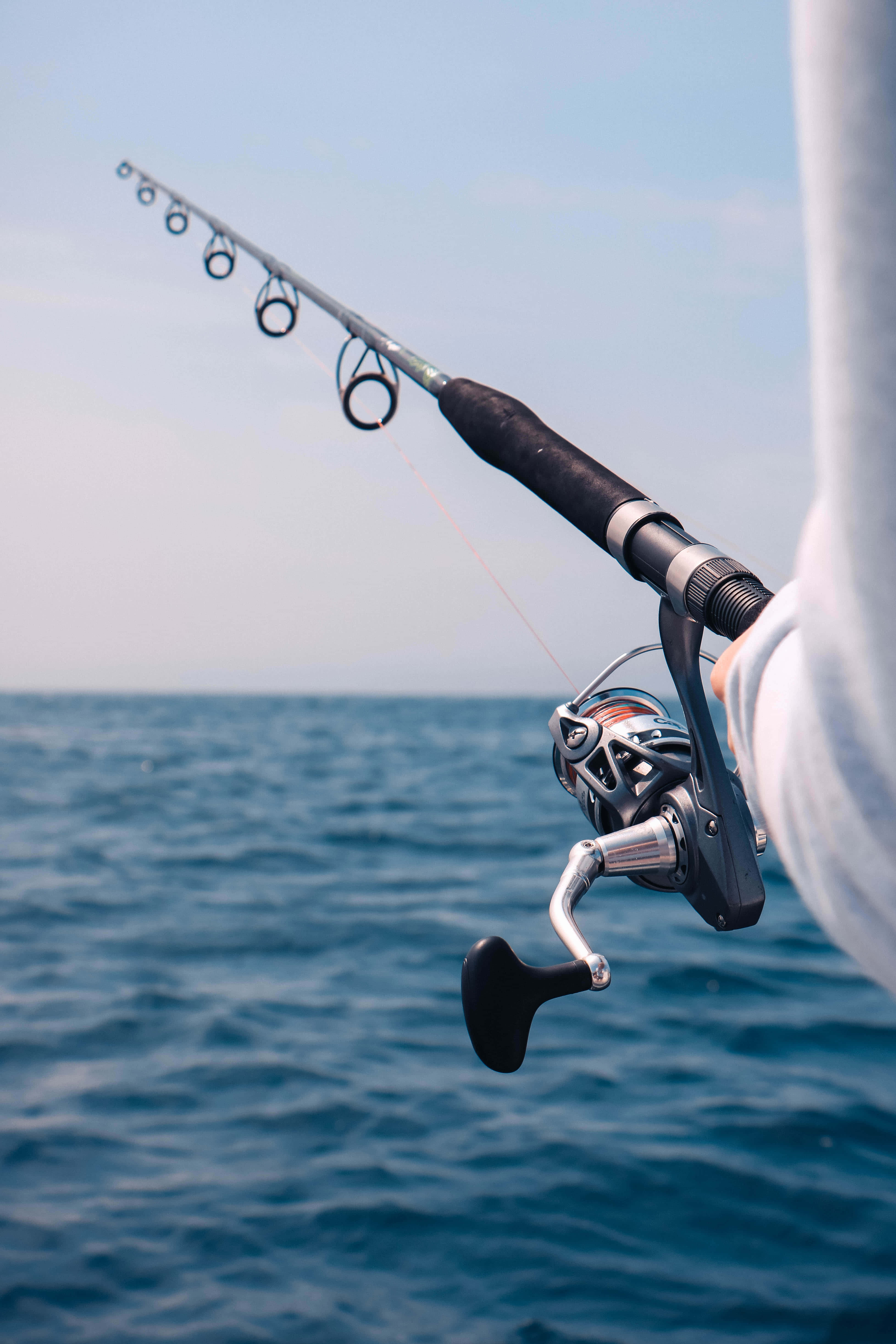 Fishing Rod 桌布 壁紙 — 特寫鏡頭：晴朗天空下，有人手持釣竿和捲線器，向開闊海洋拋投。