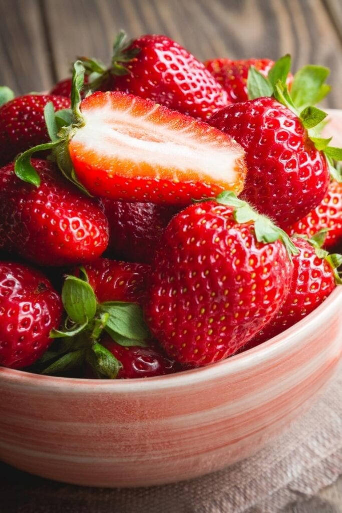 Strawberry 桌布 壁紙 — 特寫鏡頭：木質表面上的一個赤陶碗裡堆滿了飽滿的紅色草莓，頂部帶有綠色葉子。